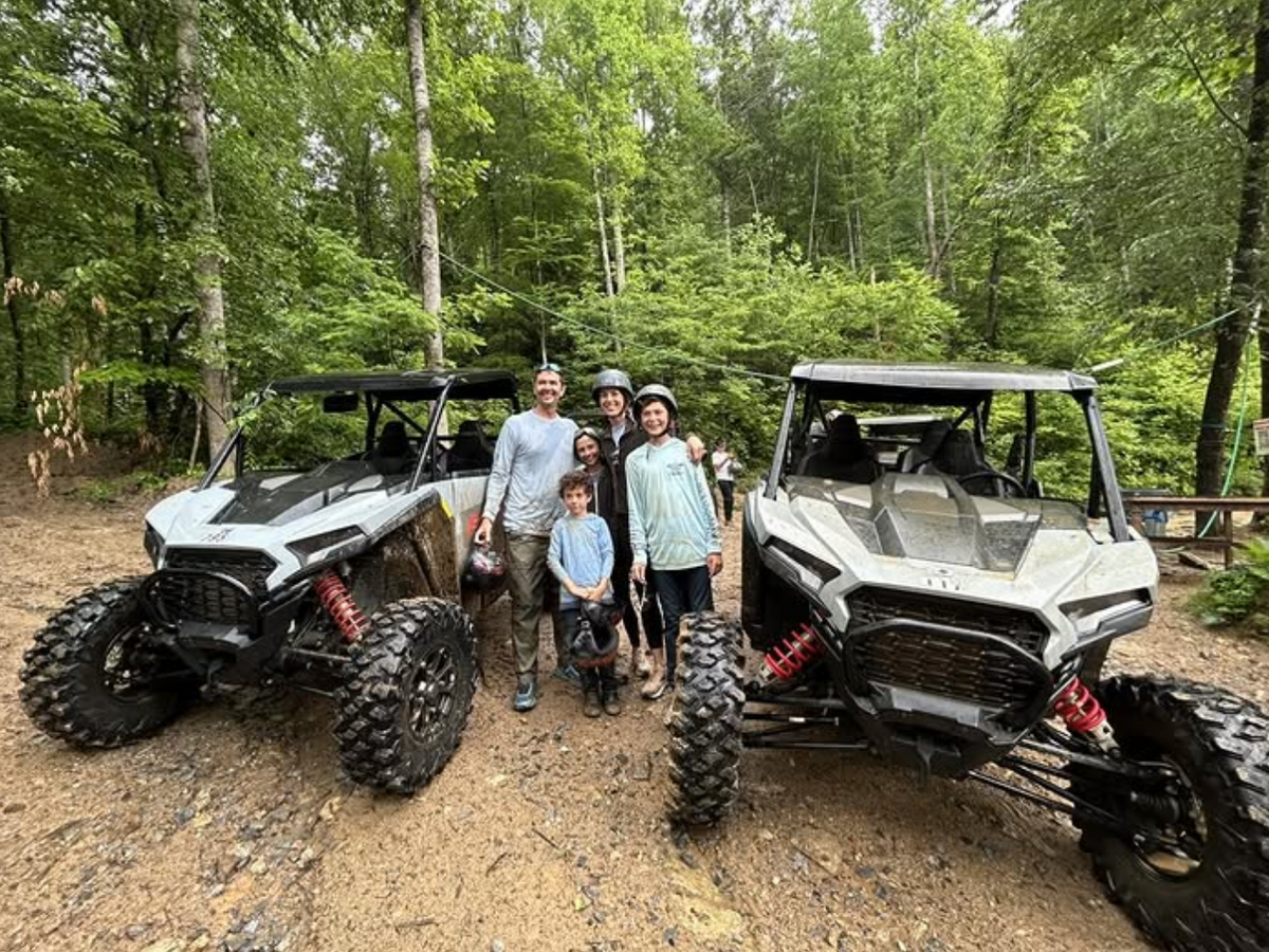 A group of six people, including two children, pose happily between two mud-covered off-road vehicles in a forested area, wearing helmets and outdoor clothing, surrounded by lush green trees and dirt trails.