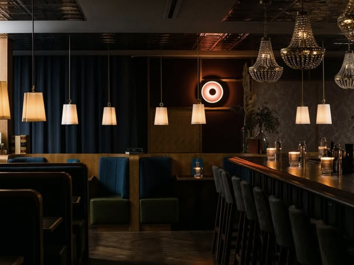 A dimly lit upscale cocktail bar with chandeliers, pendant lighting, velvet booths, and a sleek bar lined with candles.