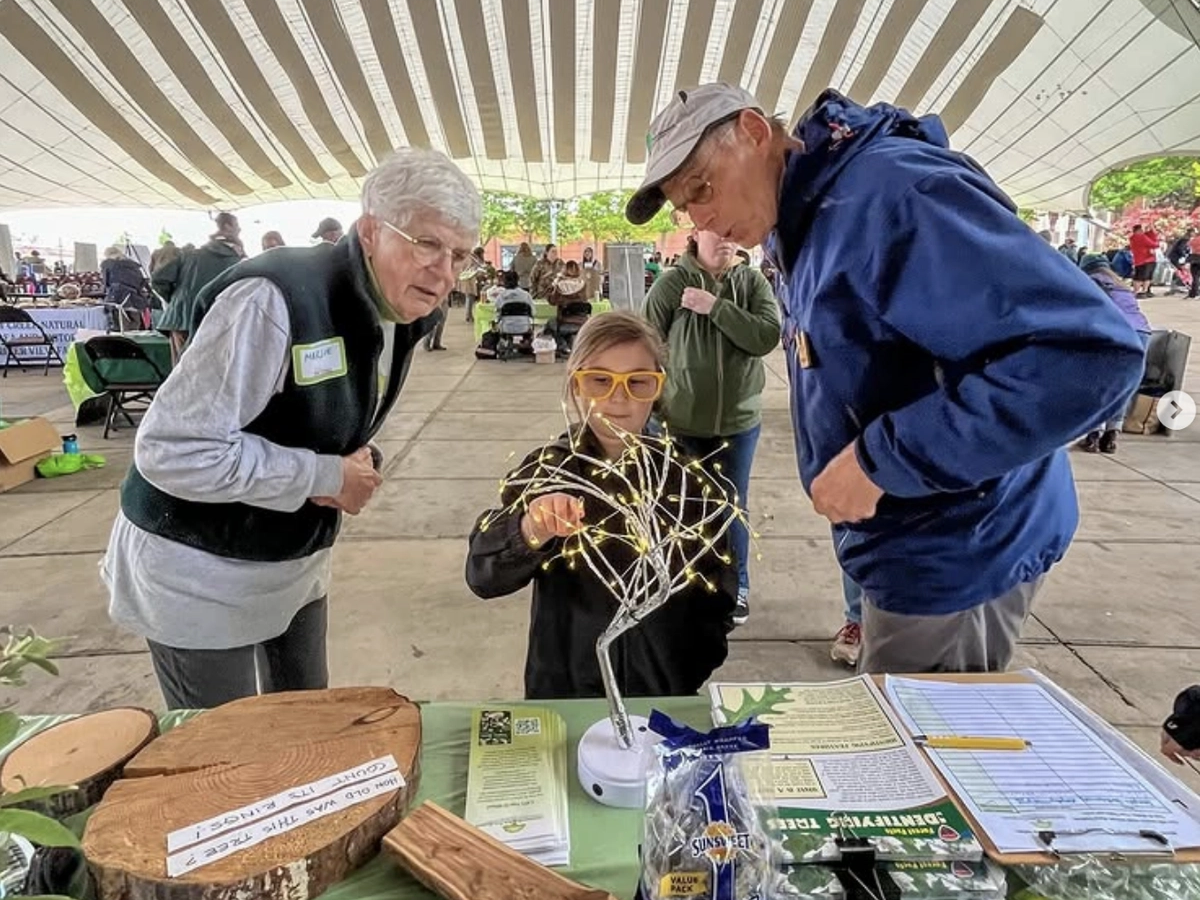 A young girl wearing yellow glasses interacts with a lit-up tree model at a booth, while two older adults look on with interest under a large event tent.