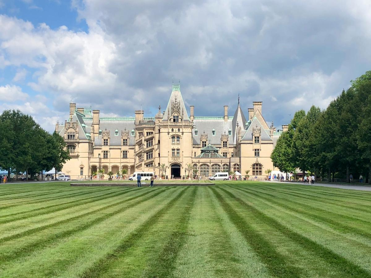 The grand Biltmore Estate in Asheville, North Carolina, stands beneath partly cloudy skies, framed by manicured green lawns and rows of trees.