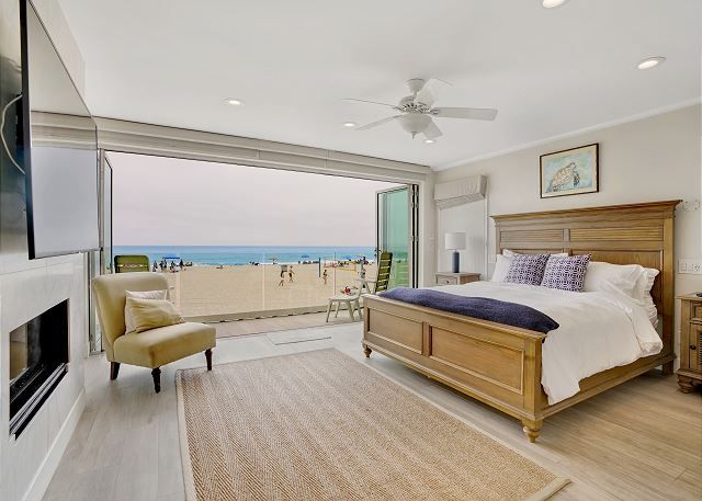 Oceanfront master bedroom in a Newport Beach vacation rental featuring a king bed, fireplace, and fully retractable glass doors opening directly to the beach.