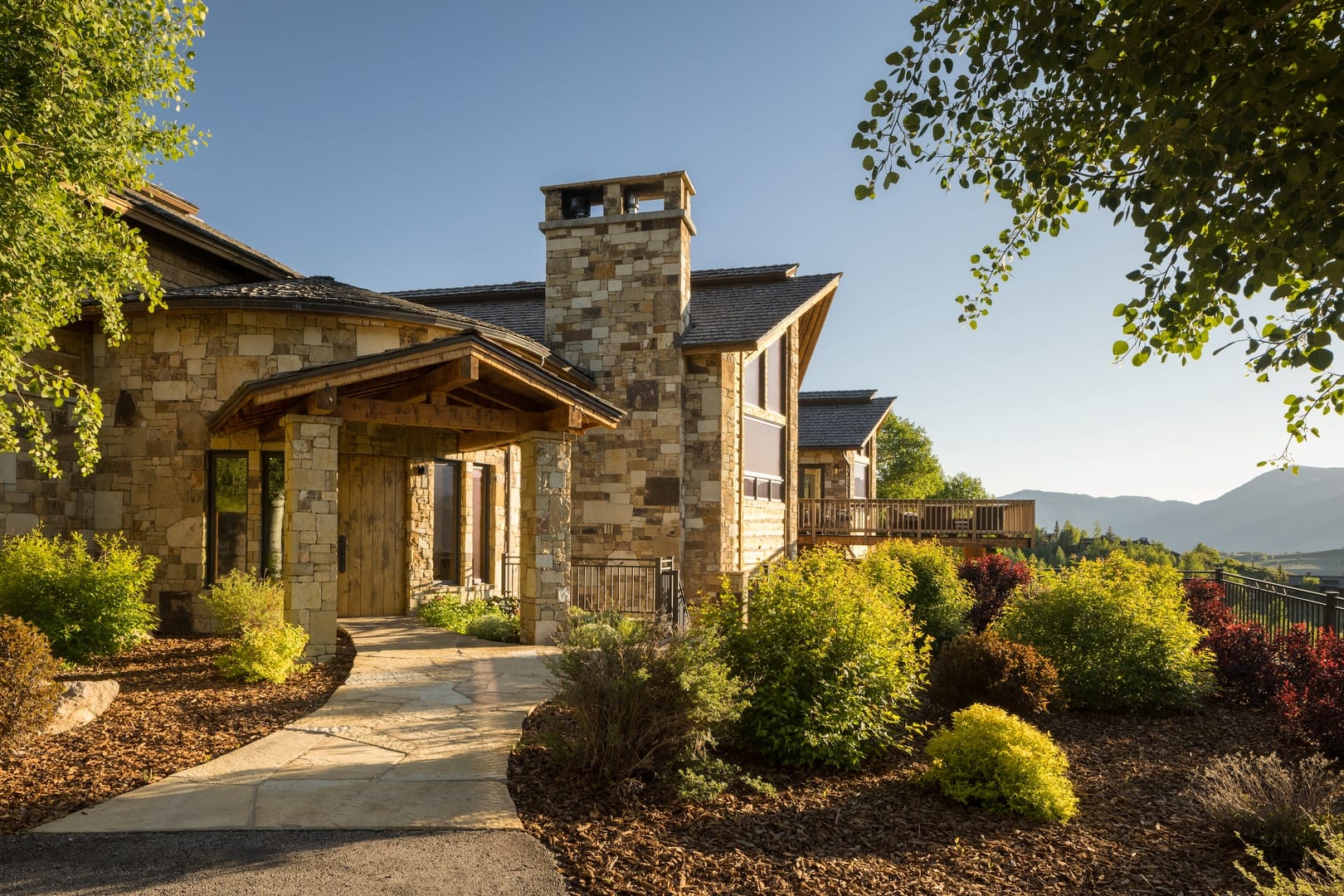 A luxurious stone mountain lodge surrounded by colorful shrubs and panoramic mountain views. The golden sunlight highlights the rustic wood and stone textures, creating a peaceful alpine escape.
