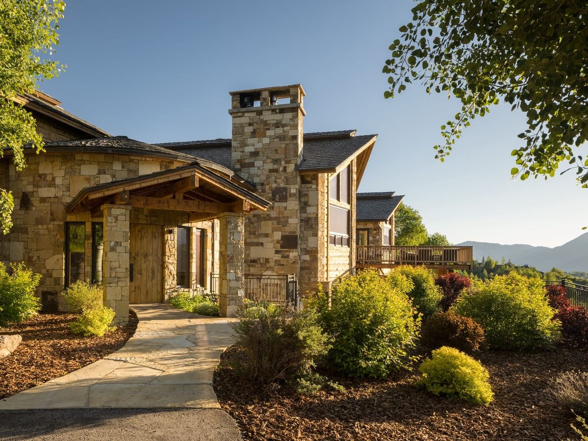 A luxurious stone mountain lodge surrounded by colorful shrubs and panoramic mountain views. The golden sunlight highlights the rustic wood and stone textures, creating a peaceful alpine escape.