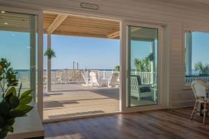 "A bright and airy beachfront home interior with large sliding glass doors opening to a spacious wooden deck. The deck features white Adirondack chairs, lounge seating, and potted plants, all overlooking the ocean. The indoor space has white shiplap walls, wood flooring, and natural light streaming in. A small sign above the door reads 'Stay Awhile,' inviting relaxation. The background reveals a clear blue sky and calm ocean waters, creating a tranquil coastal atmosphere.