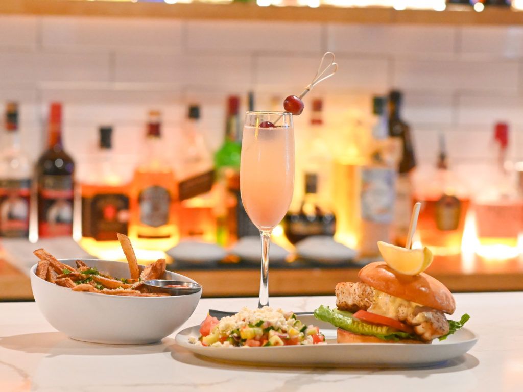 A modern dining setup showcasing crispy fries, a gourmet burger, and a refreshing cocktail against a stylish backlit bar. Perfect for a relaxed night out with upscale beach-town vibes.