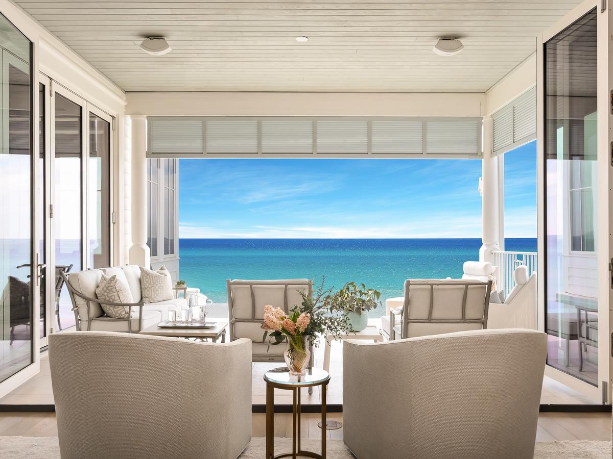 An elegant coastal living space opens onto a covered balcony with plush seating and panoramic views of the turquoise ocean under a clear blue sky.
