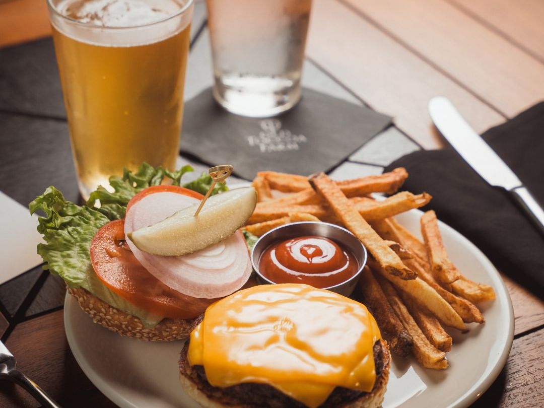 Cheeseburger with fries, ketchup, and a cold beer, all served on a wooden table. A classic all-American meal ready to be enjoyed in a relaxed setting.