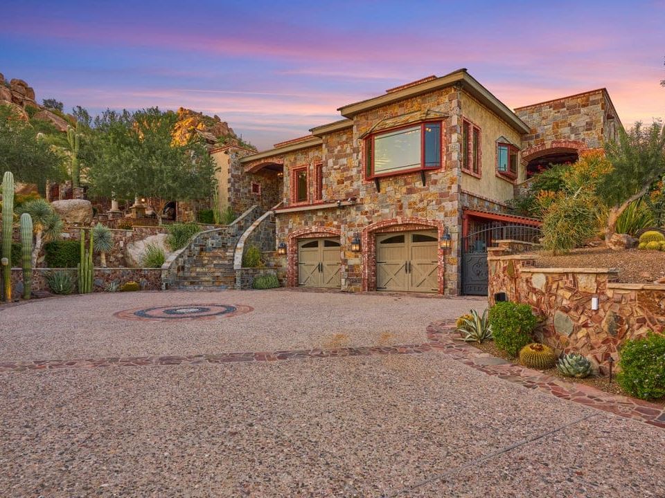 Castle on Black Mountain is a breathtaking stone estate set against desert hills with cacti and native landscaping. The home features rustic elegance with arched details, a circular drive, and scenic views of Arizona’s dramatic sunsets.