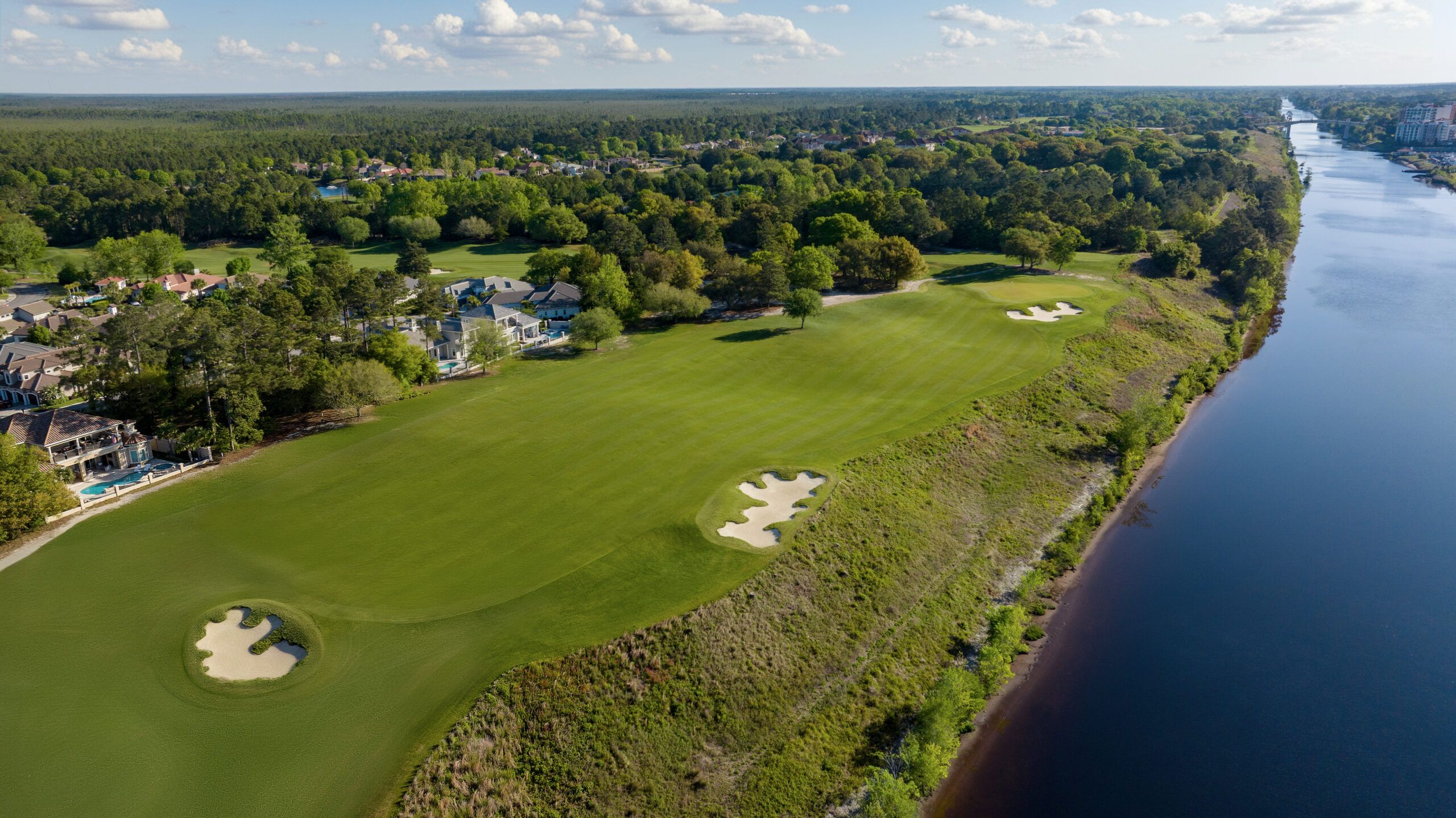 An aerial view of the Grande Dunes Resort Course in Myrtle Beach reveals manicured fairways and sand bunkers running parallel to the Intracoastal Waterway, lined by pine forest and residential homes under a partly cloudy sky. 