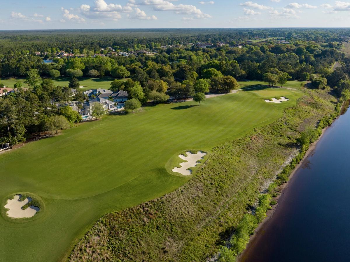 An aerial view of the Grande Dunes Resort Course in Myrtle Beach reveals manicured fairways and sand bunkers running parallel to the Intracoastal Waterway, lined by pine forest and residential homes under a partly cloudy sky.