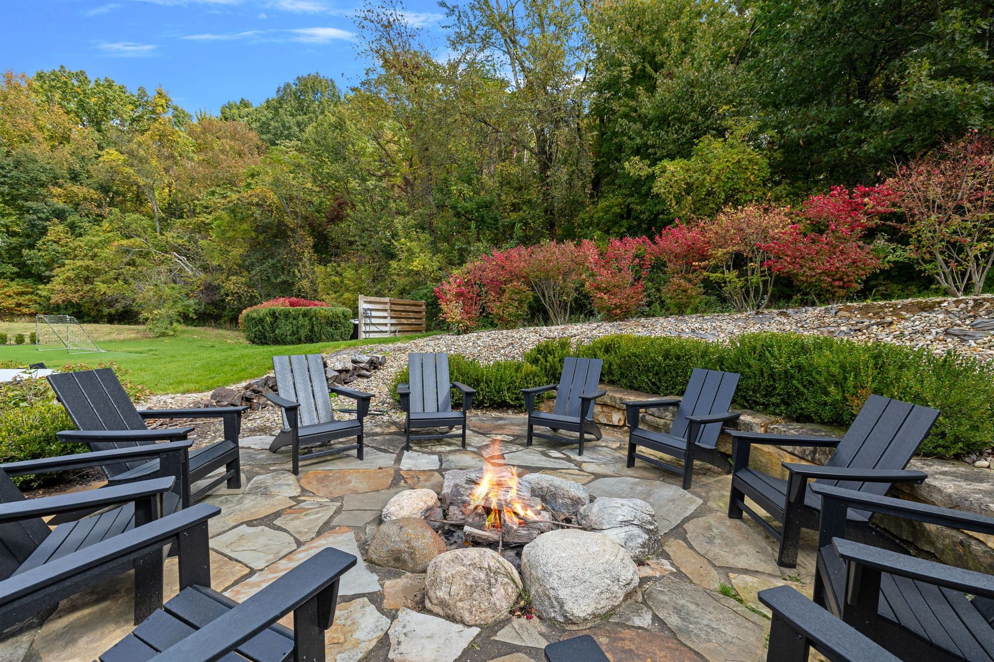Stone fire pit surrounded by Adirondack chairs, tucked into a peaceful backyard with trees and fall colors—perfect for roasting s’mores and late-night laughs with your pup by your side. A cozy outdoor spot where memories (and marshmallows) get toasted.