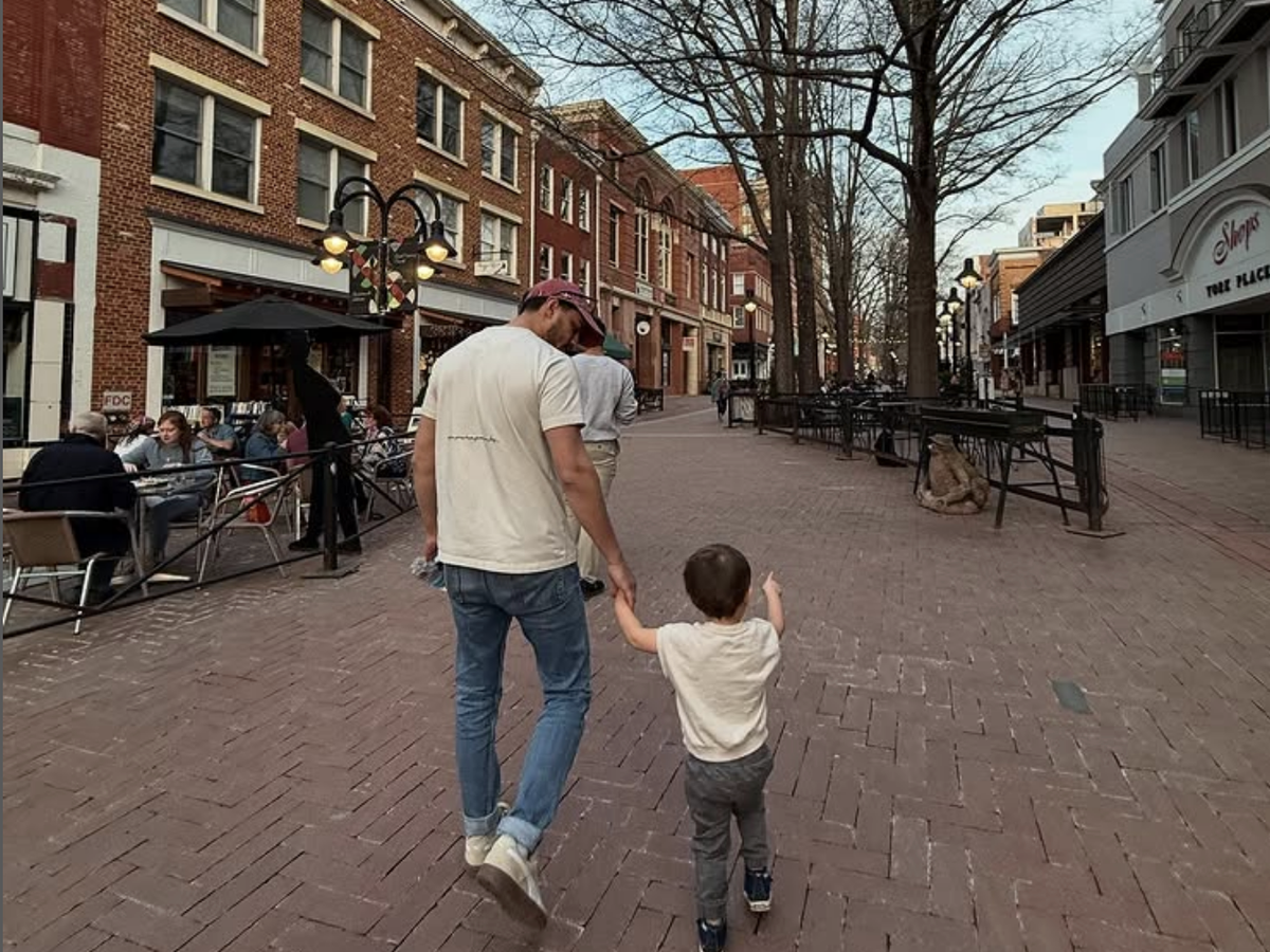 A man and a young boy walk hand-in-hand down a brick-paved pedestrian street lined with restaurants, shops, and outdoor diners on a calm afternoon.