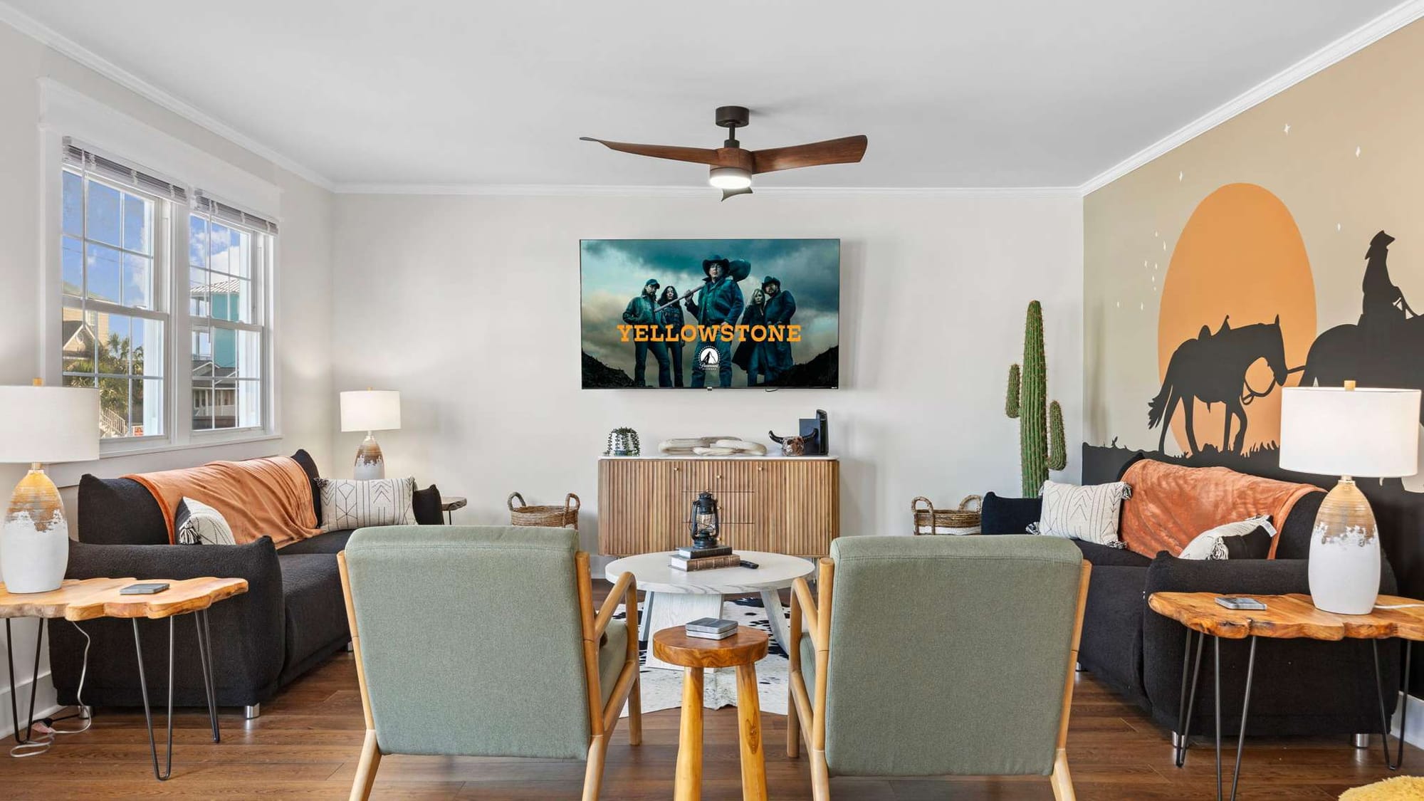 A western-inspired living room with cozy seating, rustic wood side tables, and a mural of cowboys at sunset, featuring a large TV playing Yellowstone.