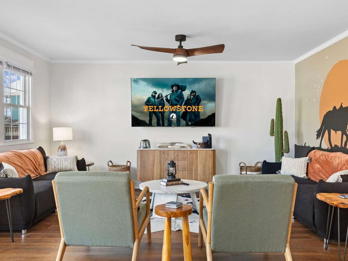 A western-inspired living room with cozy seating, rustic wood side tables, and a mural of cowboys at sunset, featuring a large TV playing Yellowstone.
