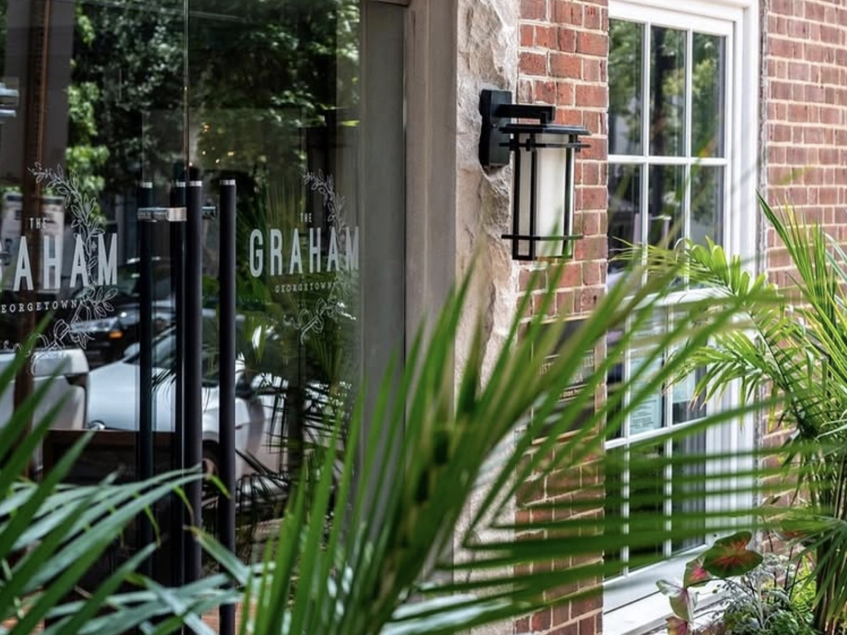 Entrance to The Graham Georgetown hotel with glass doors, brick exterior, and lush green plants framing the entryway.