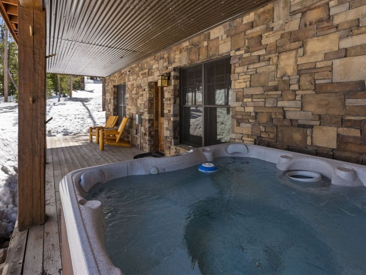 A steaming hot tub sits on a covered wooden deck beside a stone cabin wall, with snowy trees and a pair of rustic wooden chairs in the background.