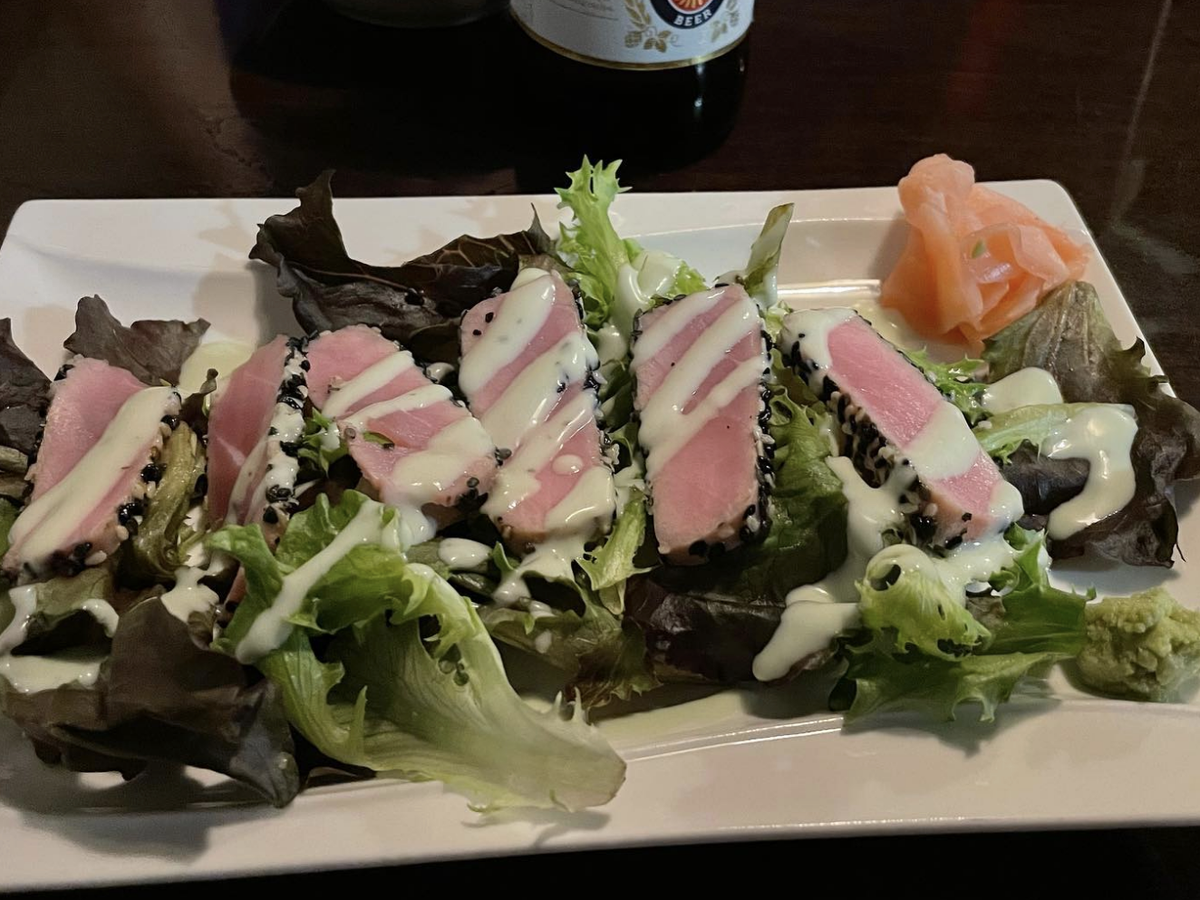 Slices of seared ahi tuna with a sesame crust are served on mixed greens, drizzled with a creamy dressing, and accompanied by pickled ginger and wasabi.