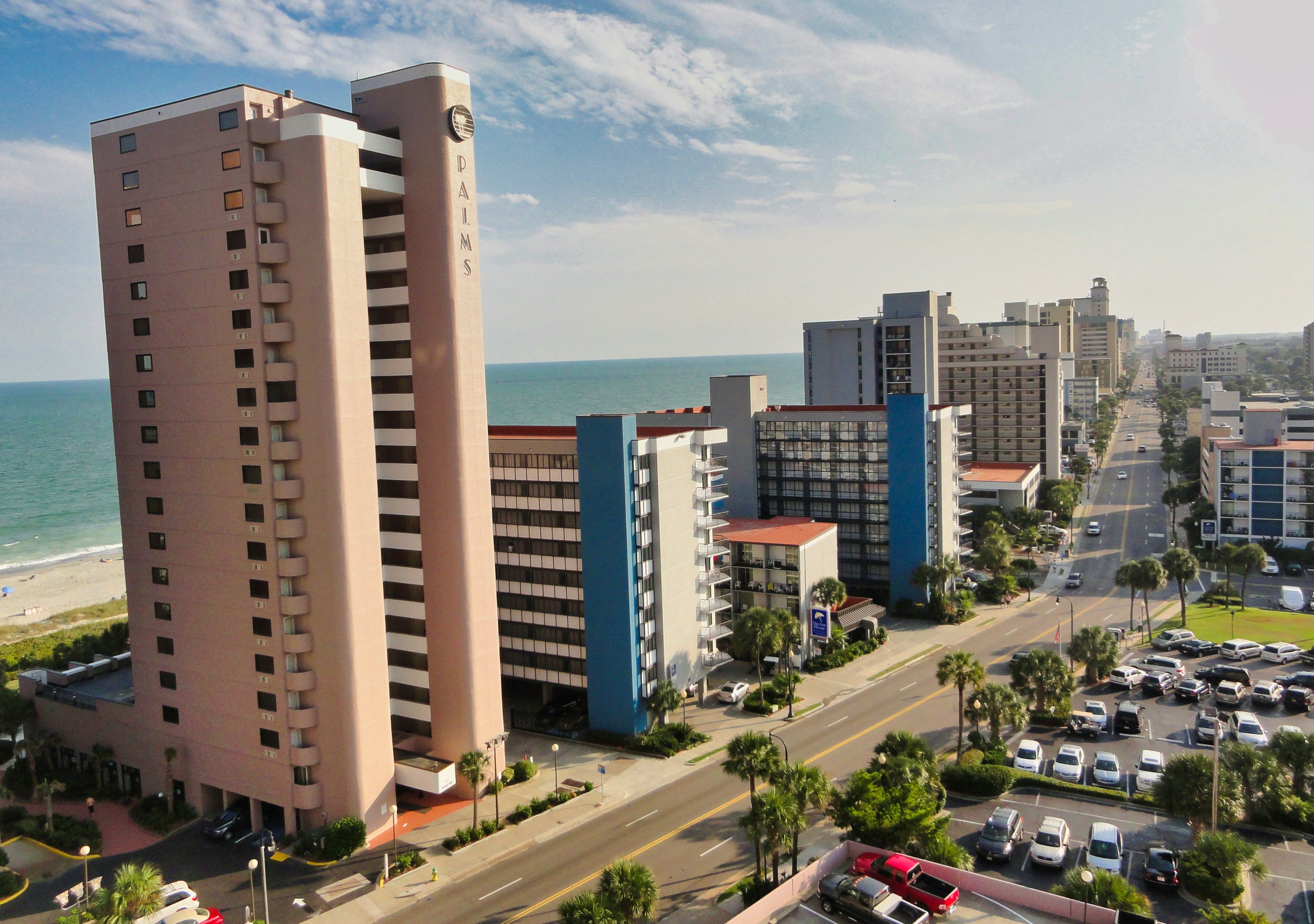 A coastal cityscape with tall beachfront condo towers lining a palm-tree-filled avenue, the ocean stretching out behind the buildings under a bright, lightly clouded sky.