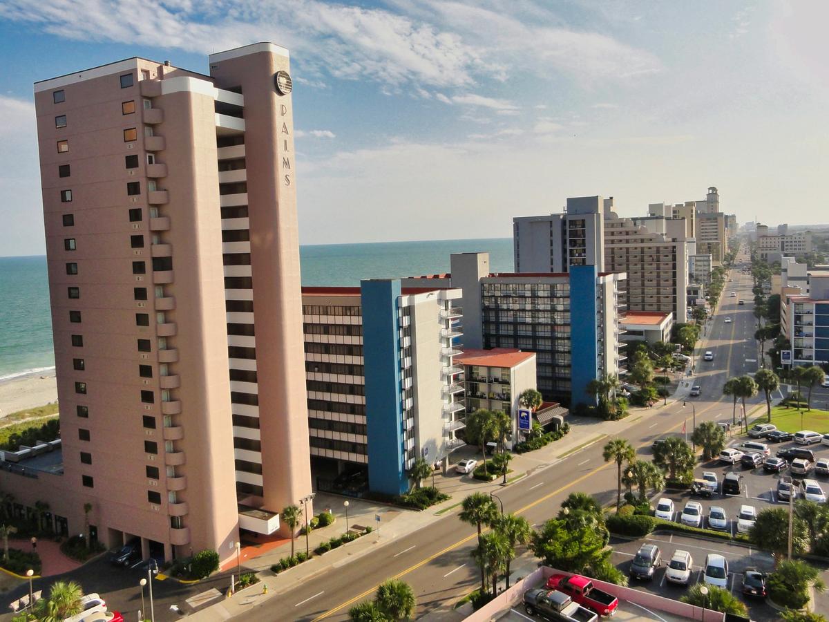 A coastal cityscape with tall beachfront condo towers lining a palm-tree-filled avenue, the ocean stretching out behind the buildings under a bright, lightly clouded sky.