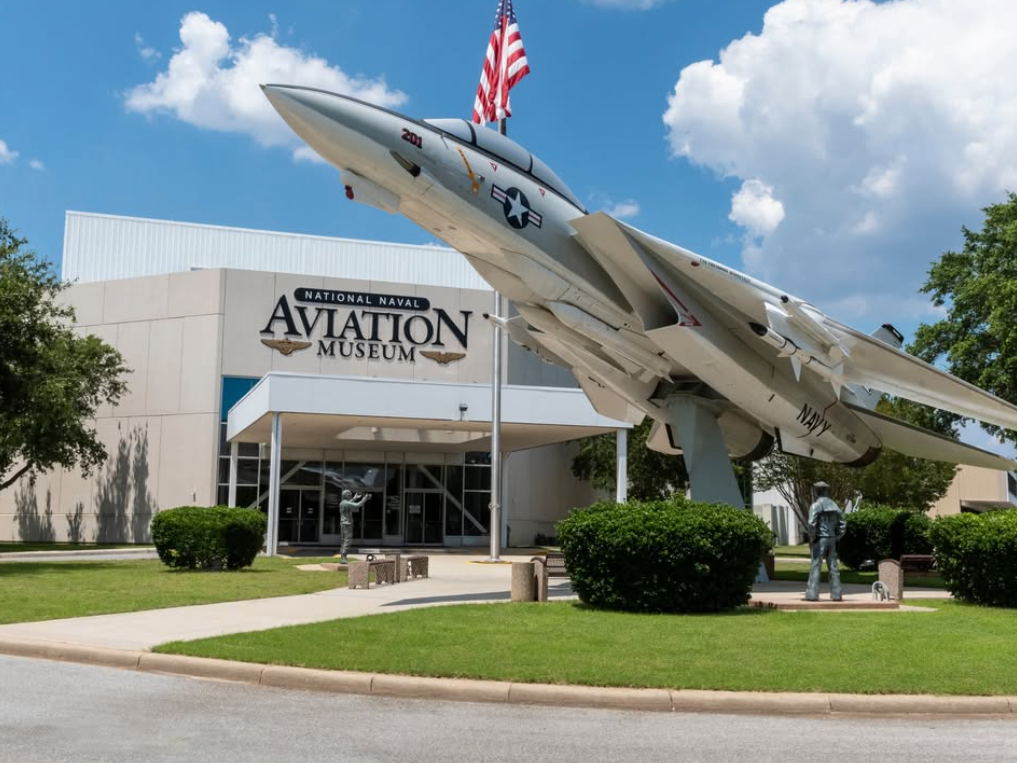 A fighter jet is mounted outside the National Naval Aviation Museum, with statues and an American flag completing the dramatic entrance