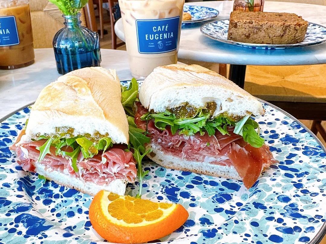 A colorful prosciutto sandwich with greens and fig spread sits on a speckled plate at Café Eugenia, paired with iced coffee and fresh flowers on the table. A cozy café perfect for brunch in style.