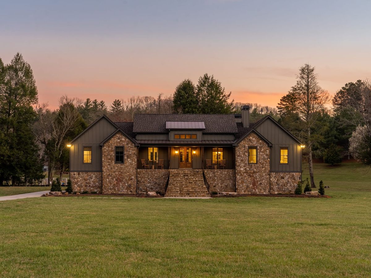 A beautiful mountain home with stone details and warm lights glowing at sunset, surrounded by open grass and tall trees. The wide front porch and peaceful setting make this retreat feel private and welcoming. It’s a perfect mountain escape for a quiet getaway in nature.