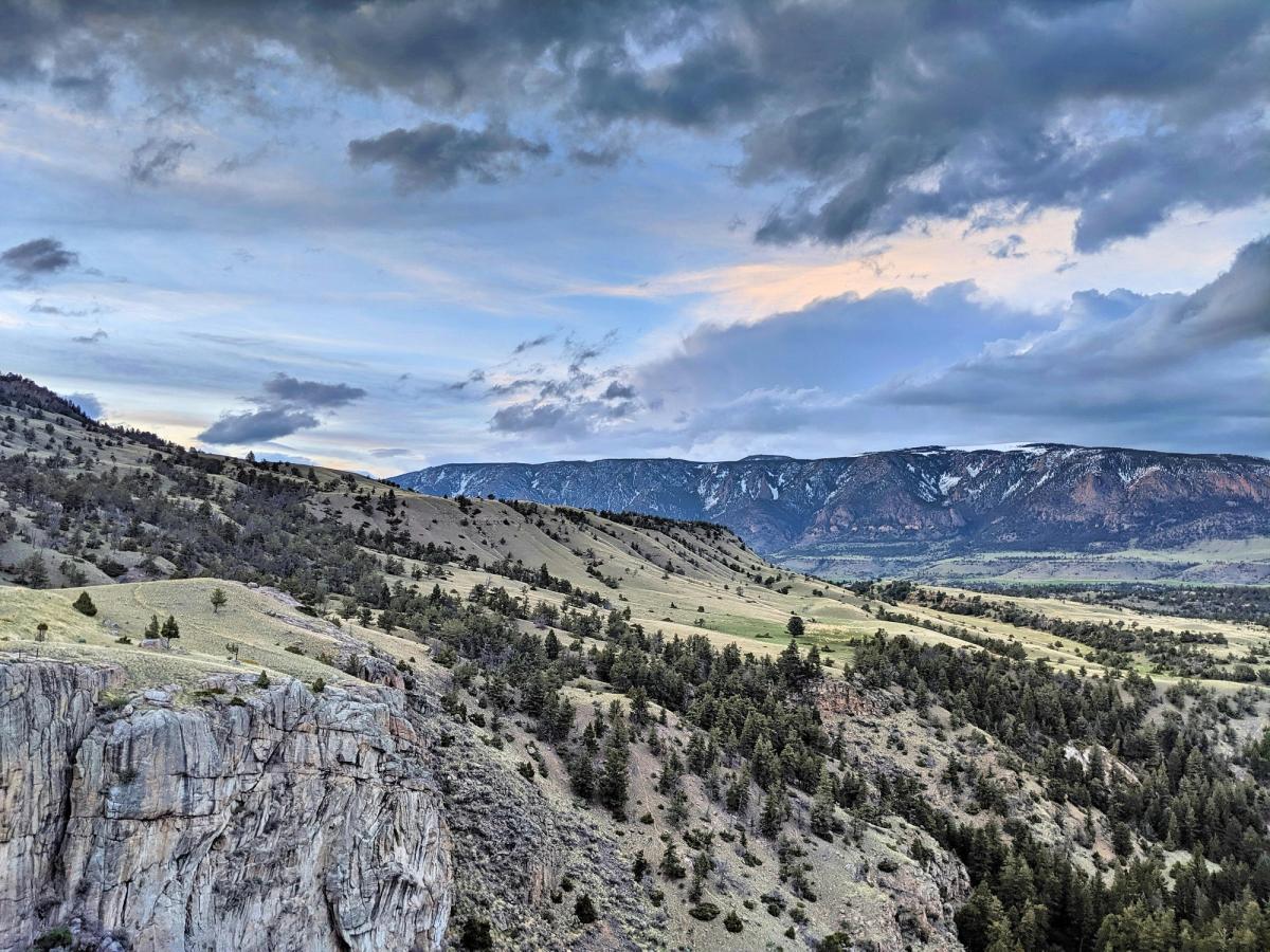 Rolling hills dotted with evergreens descend into a broad, rugged valley, framed by distant snow-streaked mountains and a dramatic sky painted in twilight hues.