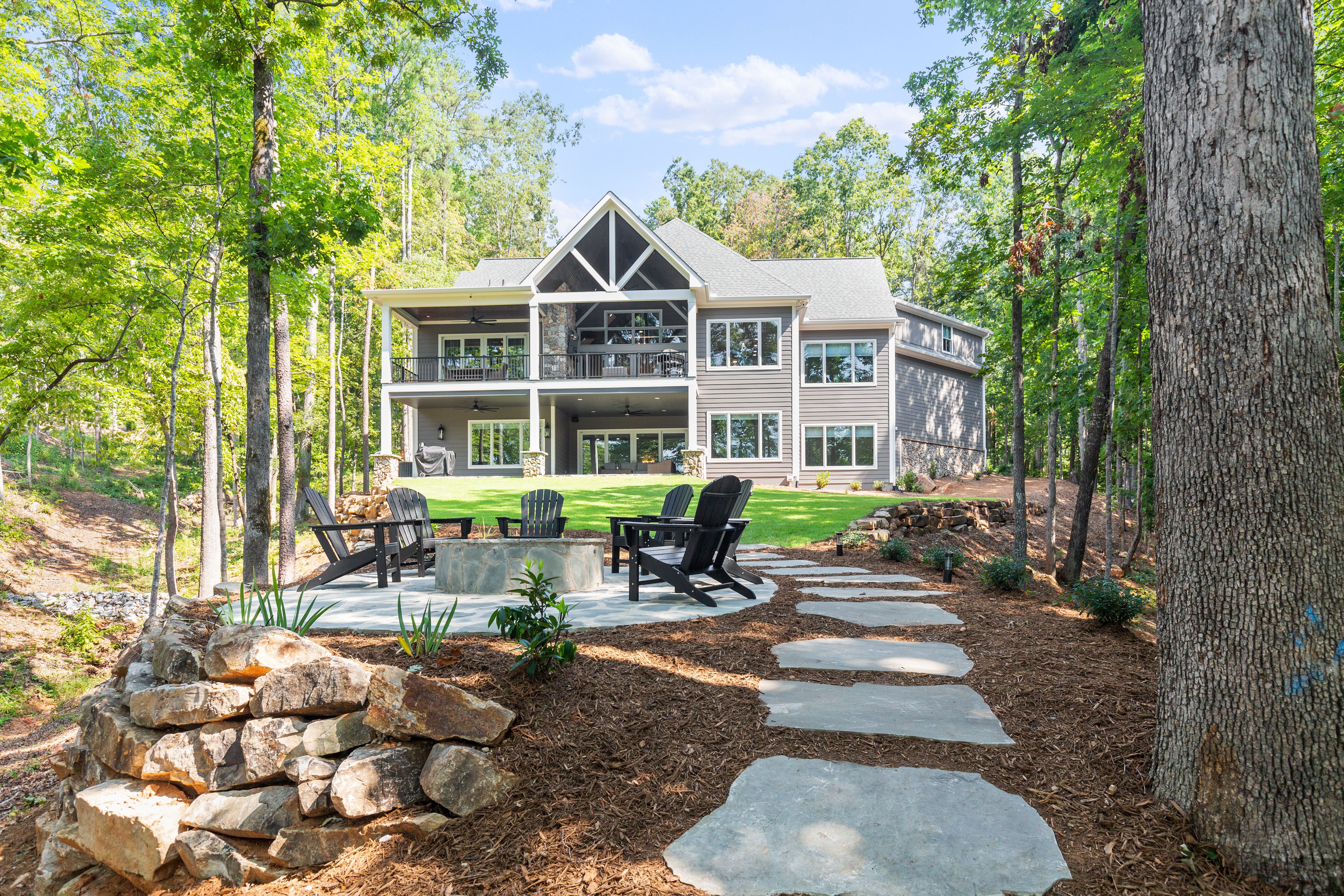 Modern lake house nestled in the woods with expansive windows, double decks, and a stone path leading to a cozy fire pit area.