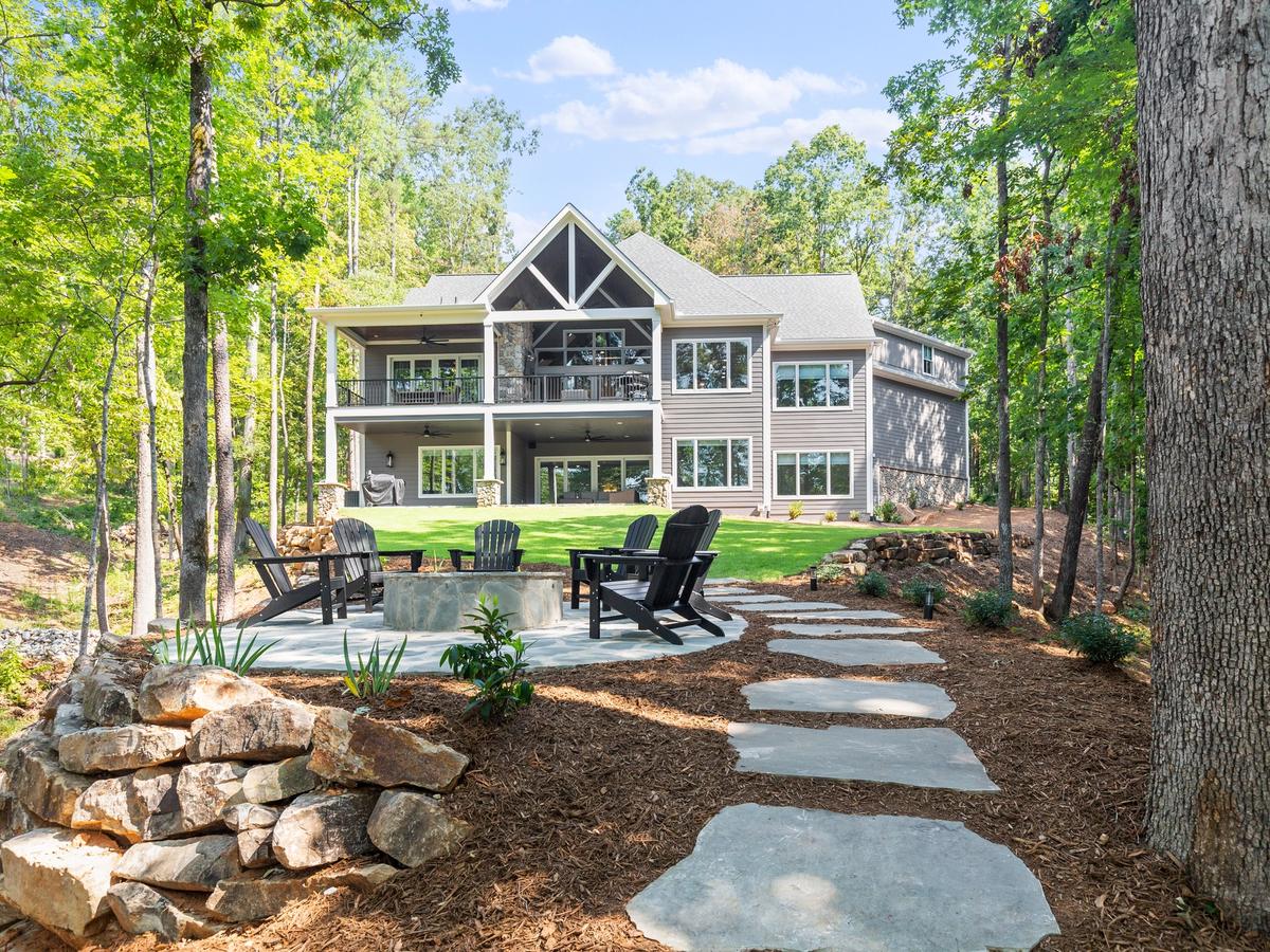 Modern lake house nestled in the woods with expansive windows, double decks, and a stone path leading to a cozy fire pit area.