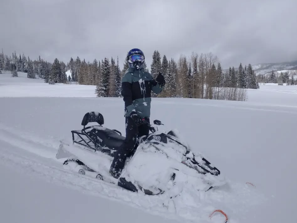 Person in snow gear and helmet gives a thumbs-up while standing on a snowmobile in a snowy, tree-lined landscape under cloudy skies.