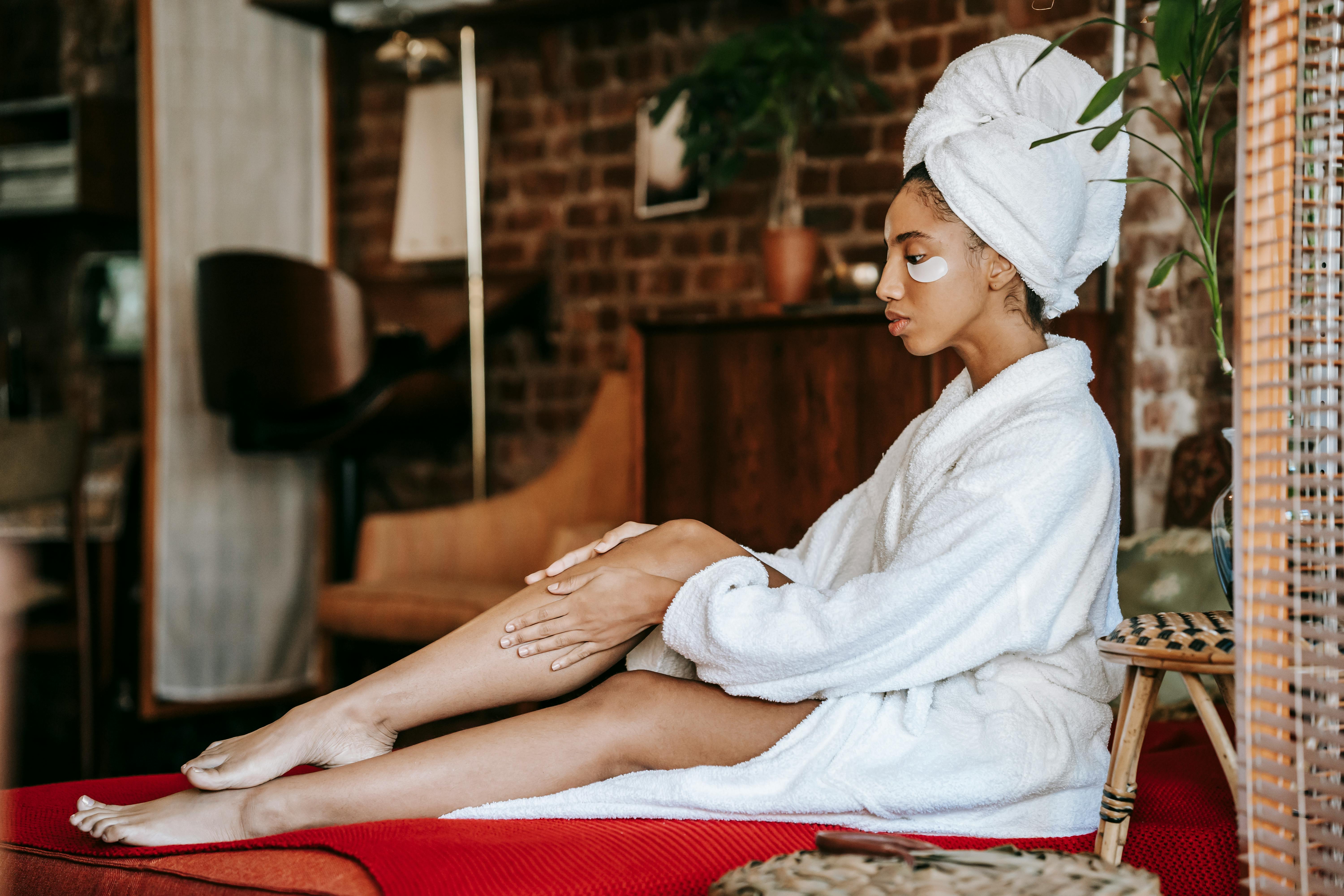 A woman in a white robe with under-eye patches sits quietly in a cozy room. The simple self-care routine highlights rest, skincare, and mindful downtime.