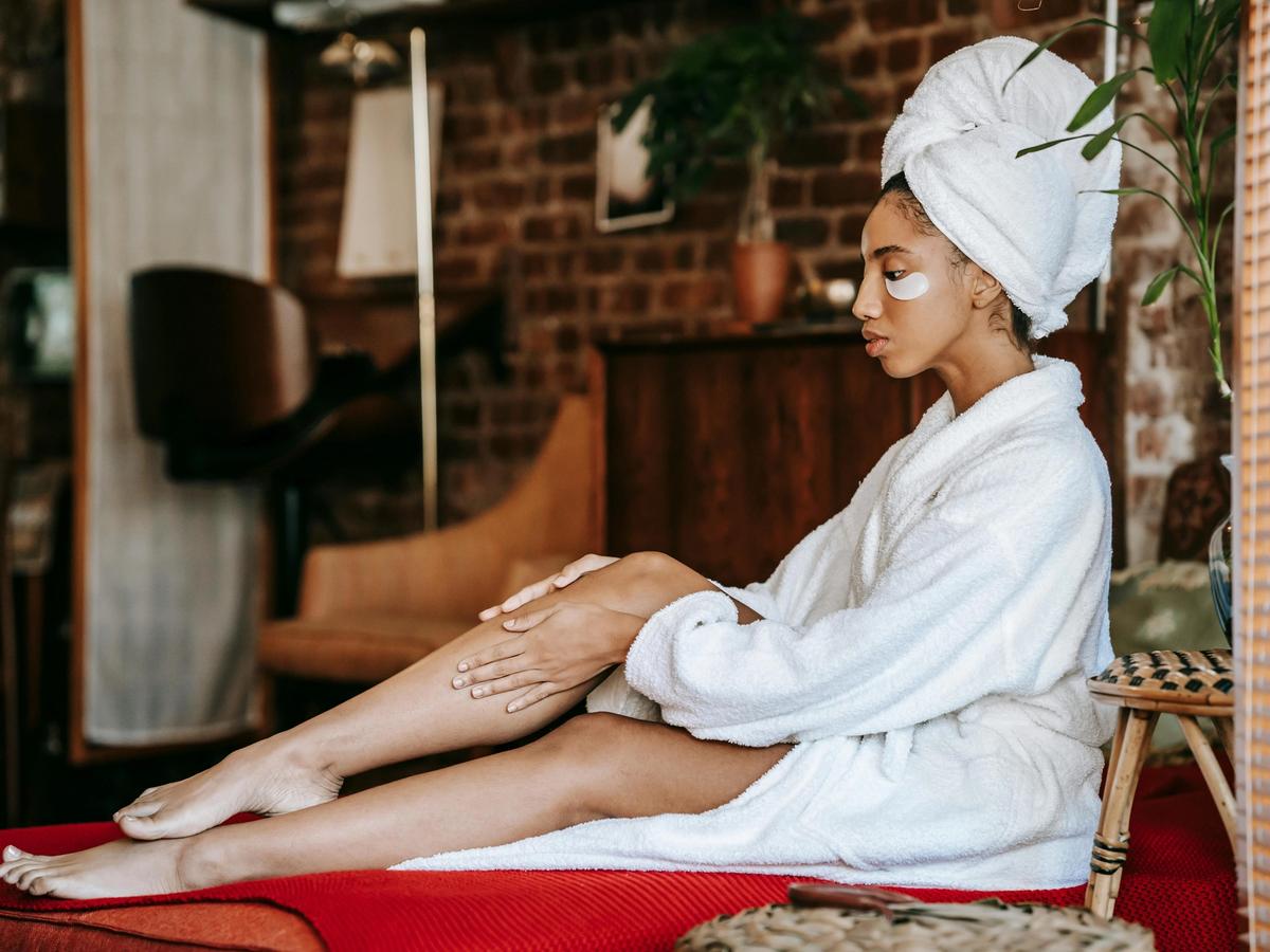 A woman in a white robe with under-eye patches sits quietly in a cozy room. The simple self-care routine highlights rest, skincare, and mindful downtime.