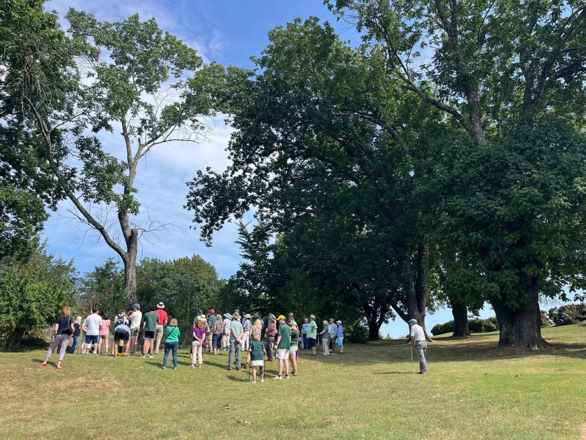 A guided tour group gathers beneath mature shade trees on the rolling lawn of James Monroe's Highland on a bright summer day. A guide leads the crowd as visitors of all ages listen among the open historic grounds.