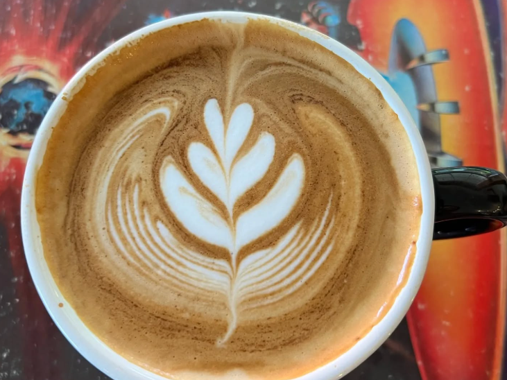 A cup of latte with a leaf-shaped design in the foam, served in a black-handled mug on a colorful table.