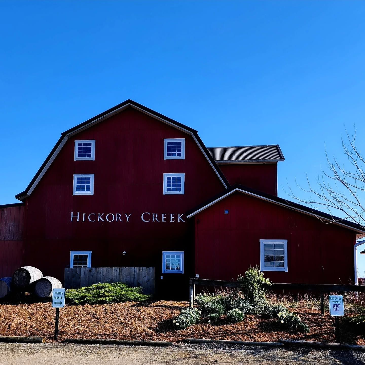 The deep red barn of Hickory Creek Winery stands against a clear blue sky, its name lettered in white across the gambrel facade. Wine barrels and a garden bed with visitor parking signs sit in the foreground on a crisp, sunny day.