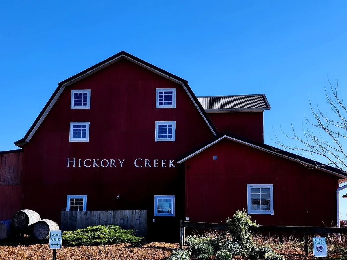 The deep red barn of Hickory Creek Winery stands against a clear blue sky, its name lettered in white across the gambrel facade. Wine barrels and a garden bed with visitor parking signs sit in the foreground on a crisp, sunny day.