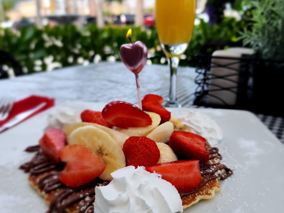 A decadent breakfast crepe topped with strawberries, bananas, whipped cream, and drizzled chocolate, paired with a mimosa. It’s a sweet Parisian-inspired escape in the sunshine