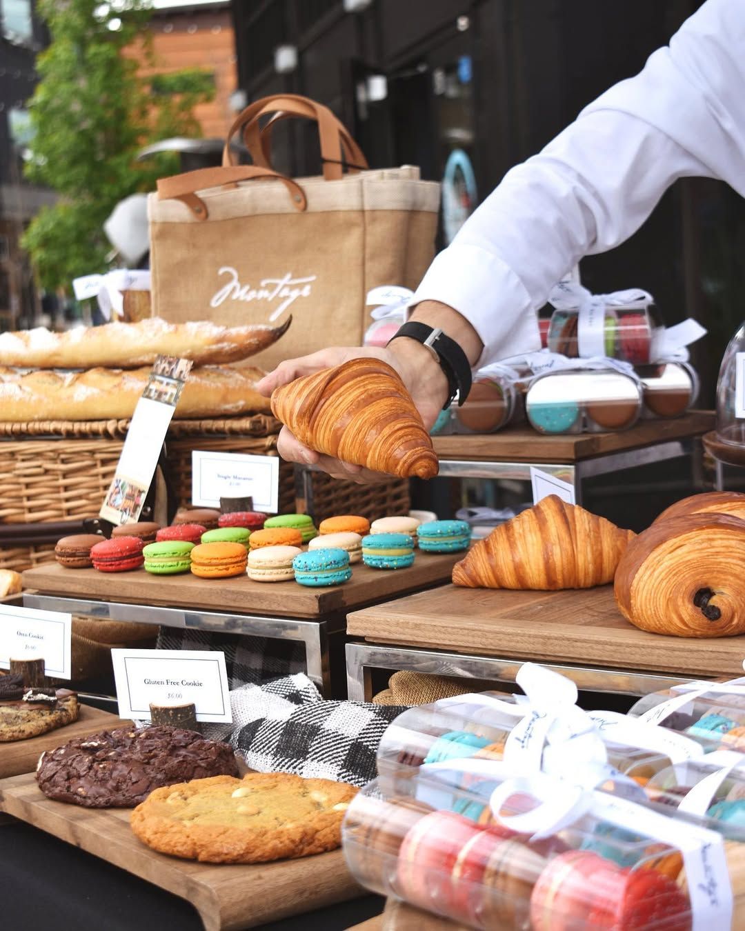 A vendor arranges fresh croissants, colorful macarons, and artisan pastries at an outdoor market stall. The inviting setup and attention to detail showcase the charm of local Montana flavors.