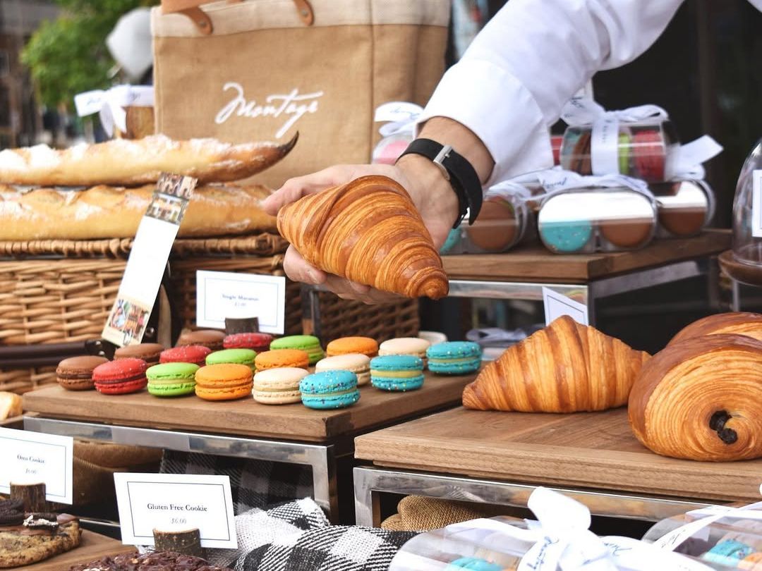 A vendor arranges fresh croissants, colorful macarons, and artisan pastries at an outdoor market stall. The inviting setup and attention to detail showcase the charm of local Montana flavors.