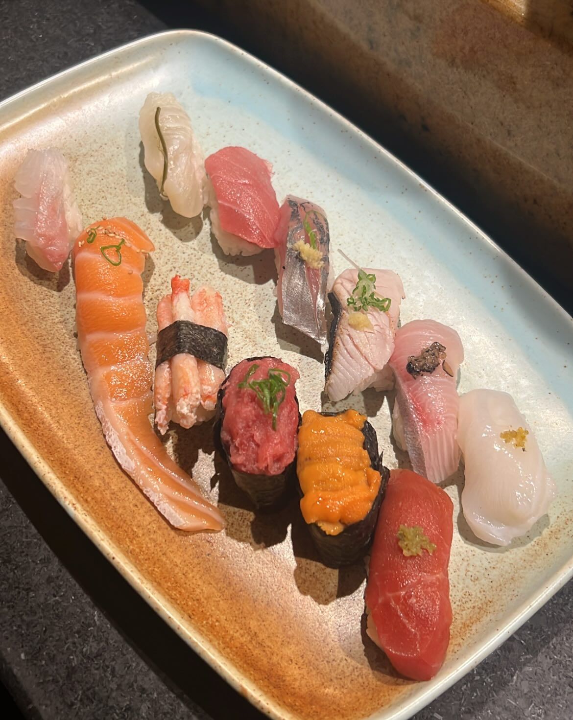 An assortment of fresh nigiri and gunkan sushi fans across a speckled ceramic platter, featuring salmon, tuna, yellowtail, uni, and other pristine cuts of fish. Sunlight highlights the vibrant colors and delicate garnishes of the expertly prepared selection.