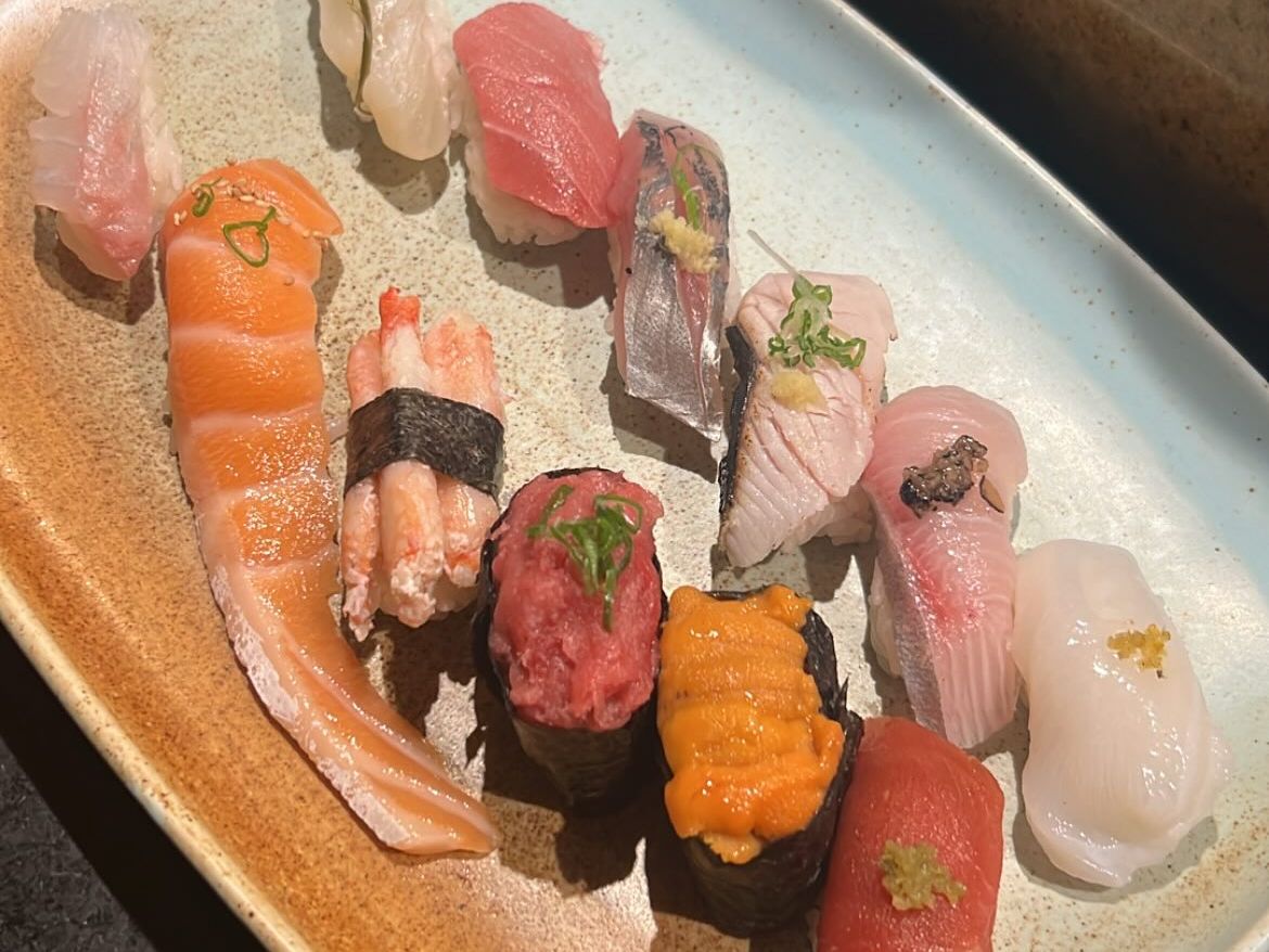 An assortment of fresh nigiri and gunkan sushi fans across a speckled ceramic platter, featuring salmon, tuna, yellowtail, uni, and other pristine cuts of fish. Sunlight highlights the vibrant colors and delicate garnishes of the expertly prepared selection.