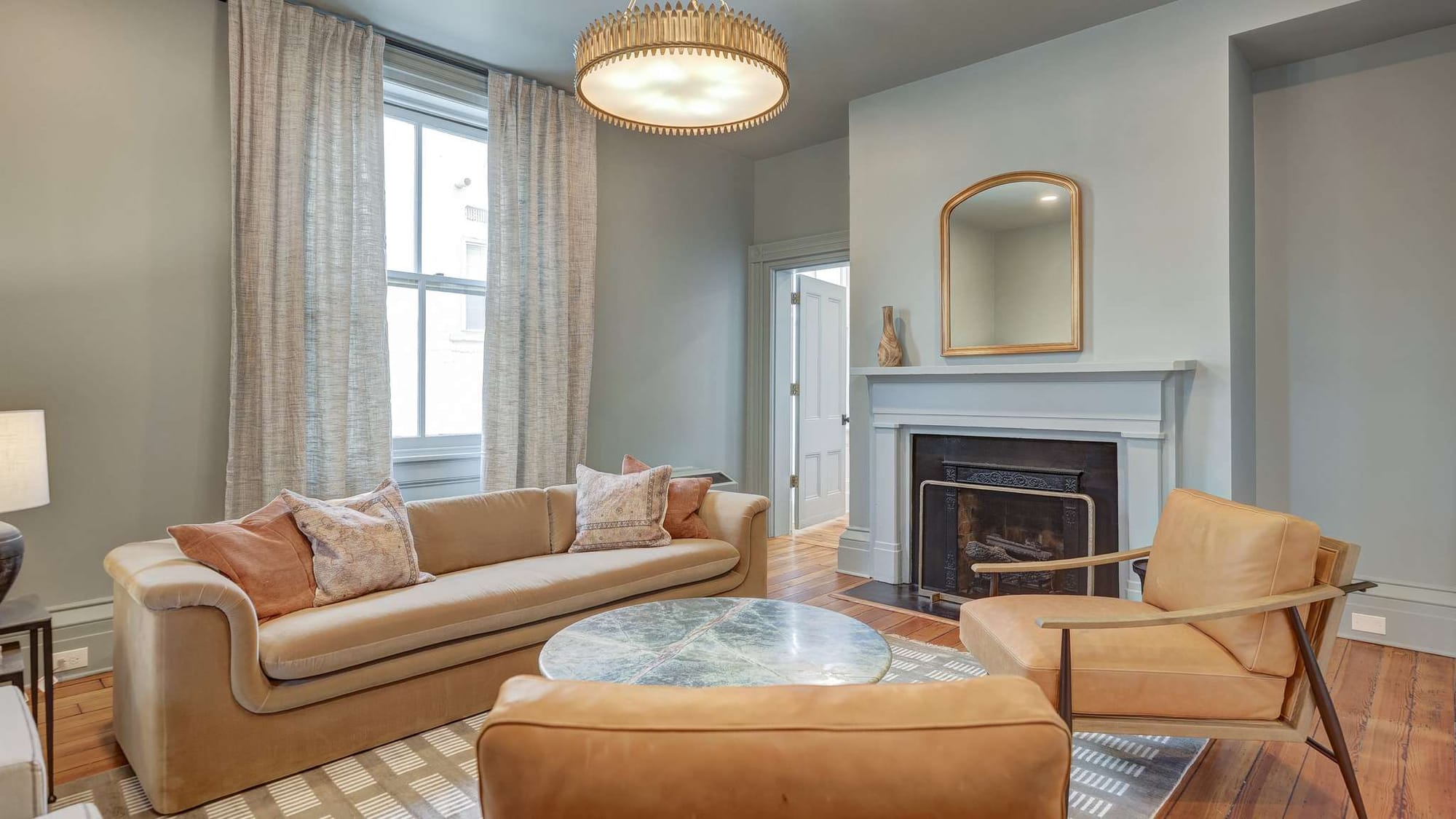 A sunlit sitting room at 200 South Street Inn features a camel-toned sofa and leather chairs arranged around a marble-top coffee table, with a white-mantled fireplace as the focal point. Sage-gray walls, a gold chandelier, and tall windows with linen drapes give the room a relaxed, refined character.