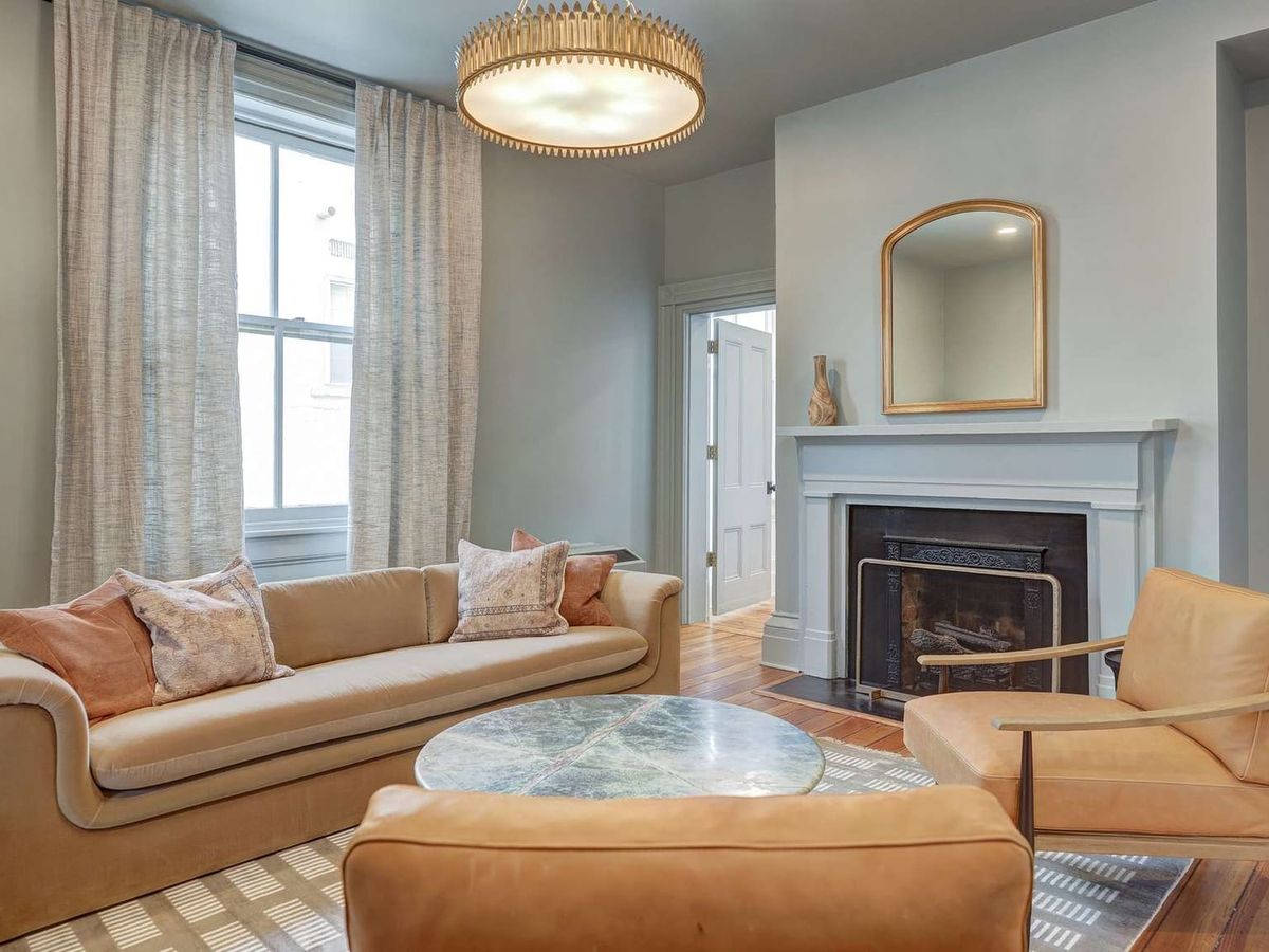 A sunlit sitting room at 200 South Street Inn features a camel-toned sofa and leather chairs arranged around a marble-top coffee table, with a white-mantled fireplace as the focal point. Sage-gray walls, a gold chandelier, and tall windows with linen drapes give the room a relaxed, refined character.