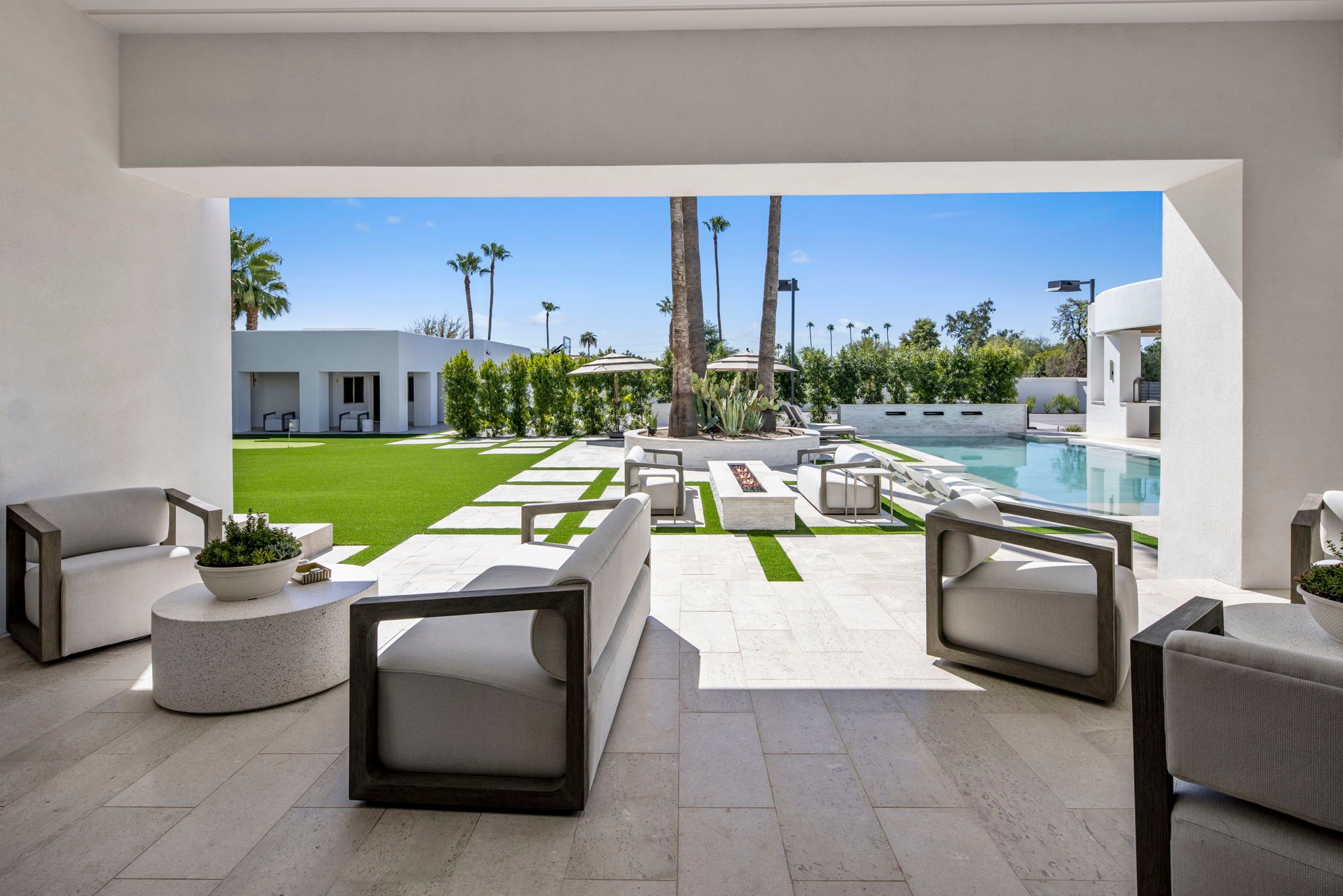 A modern luxury desert home patio with minimalist seating, a geometric lawn, fire feature, and pool framed by palm trees and clean white architecture.