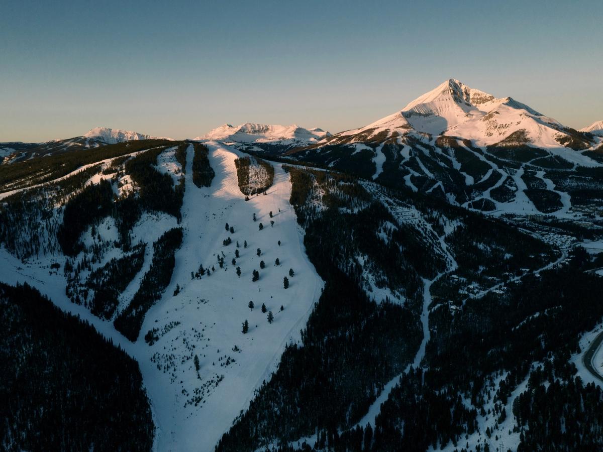 Snow-covered ski slopes wind through forested mountains at sunset, with a sharp peak illuminated in the background.