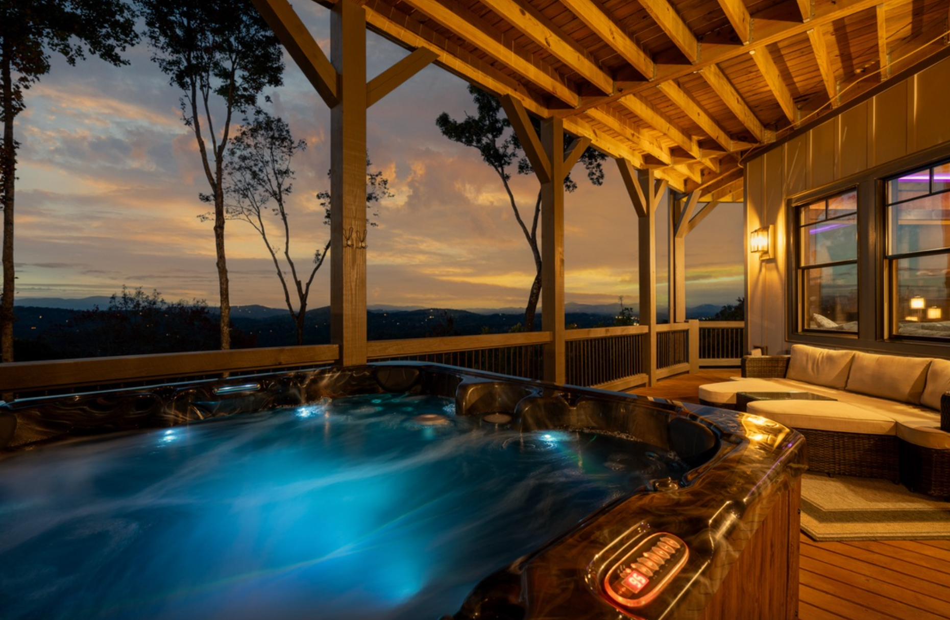 Covered mountain-view porch at sunset featuring a glowing hot tub, warm wood beams, and a cozy outdoor seating area overlooking rolling hills.