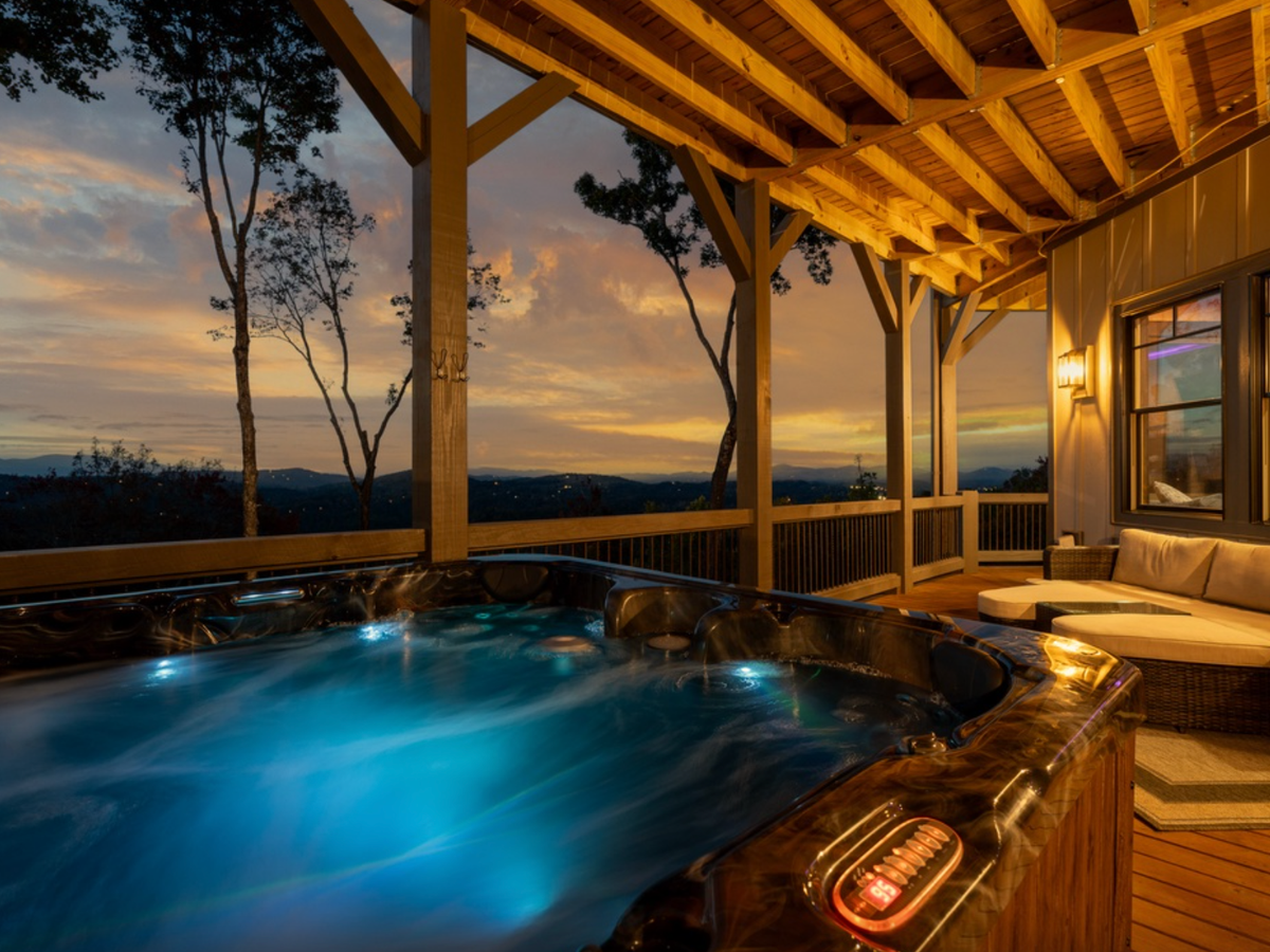 Covered mountain-view porch at sunset featuring a glowing hot tub, warm wood beams, and a cozy outdoor seating area overlooking rolling hills.