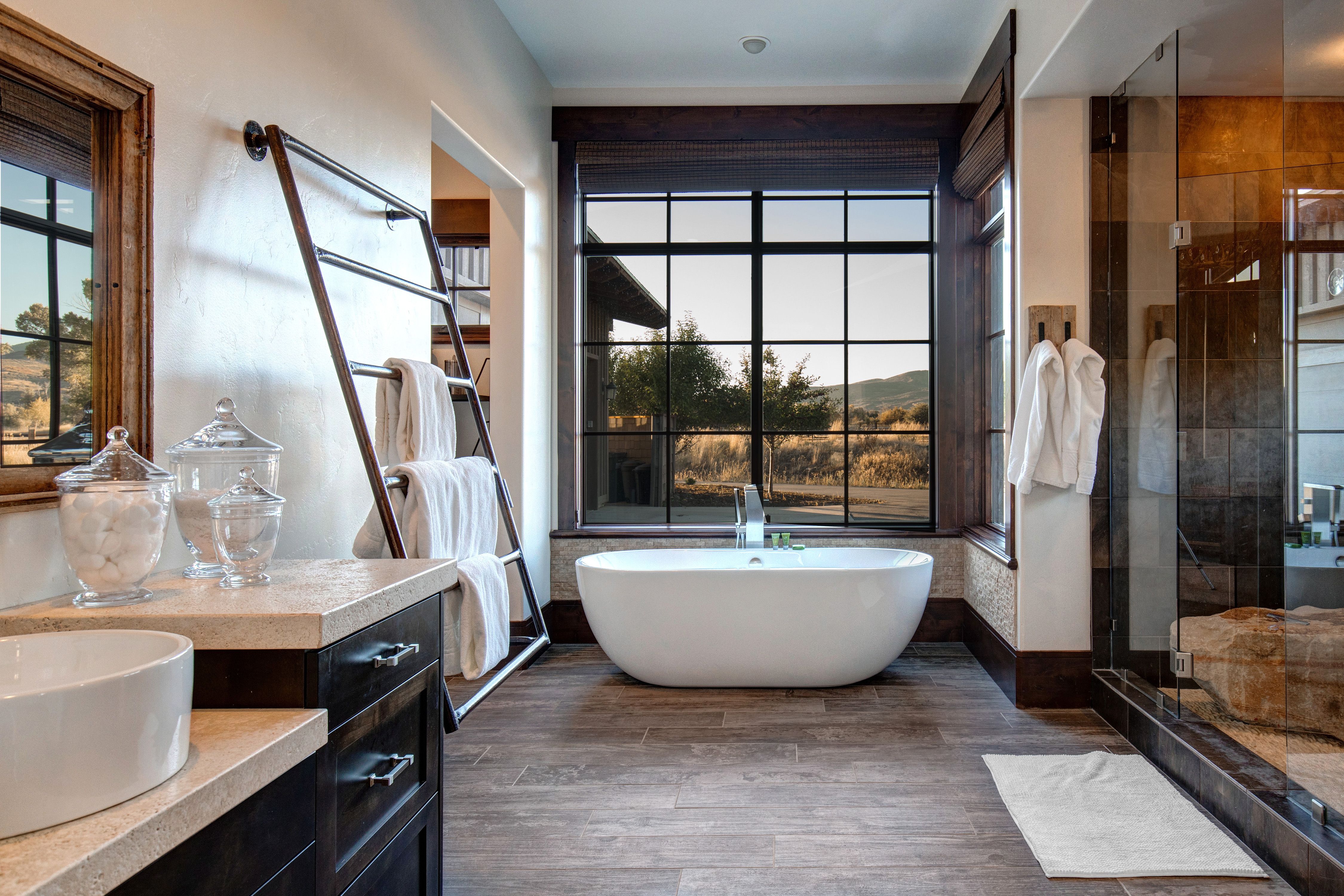A serene bathroom featuring a freestanding soaking tub by a large picture window with countryside views, a glass-enclosed shower, wood-look tile floors, and neatly rolled towels on a modern metal rack.