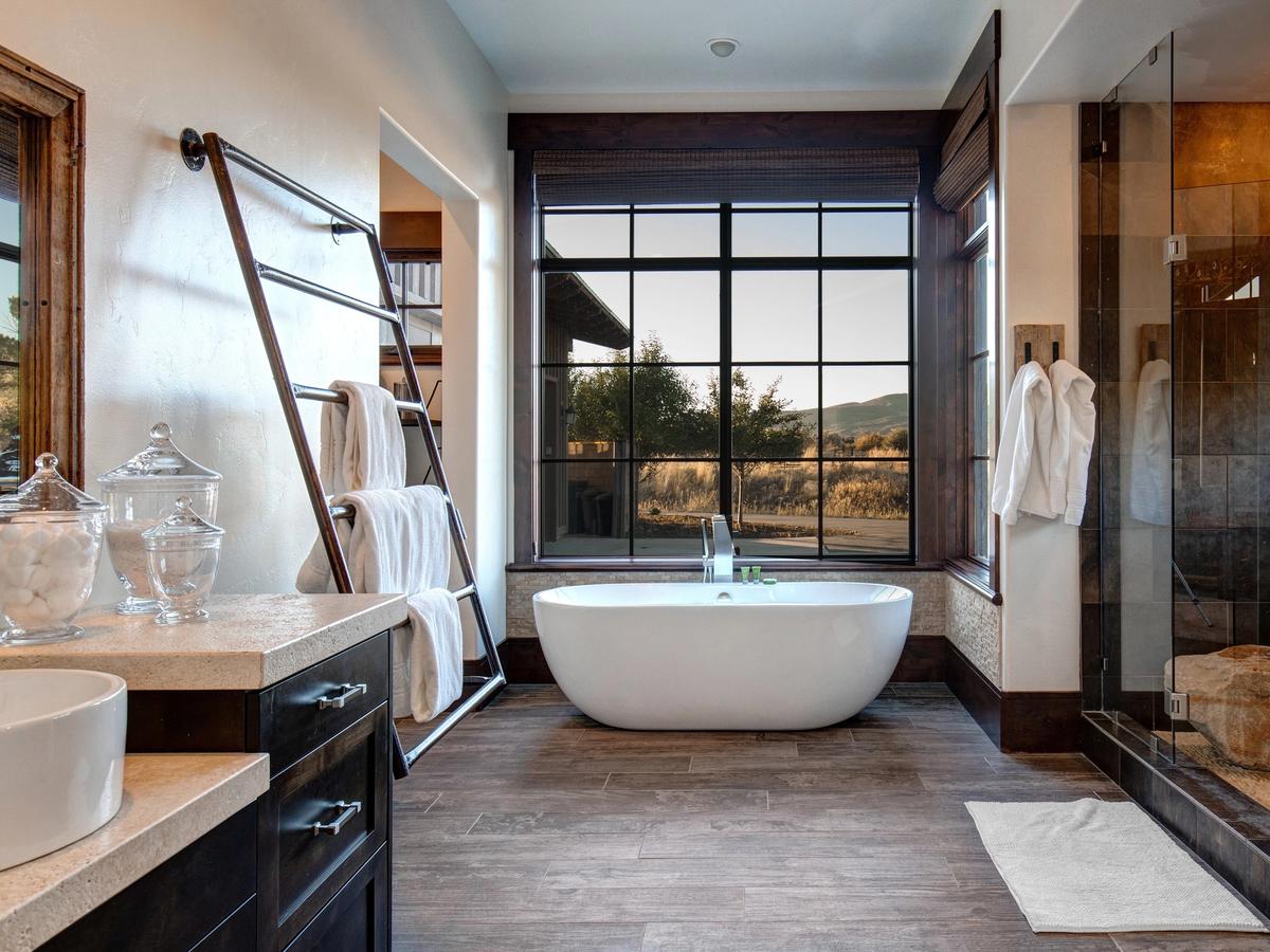A serene bathroom featuring a freestanding soaking tub by a large picture window with countryside views, a glass-enclosed shower, wood-look tile floors, and neatly rolled towels on a modern metal rack.