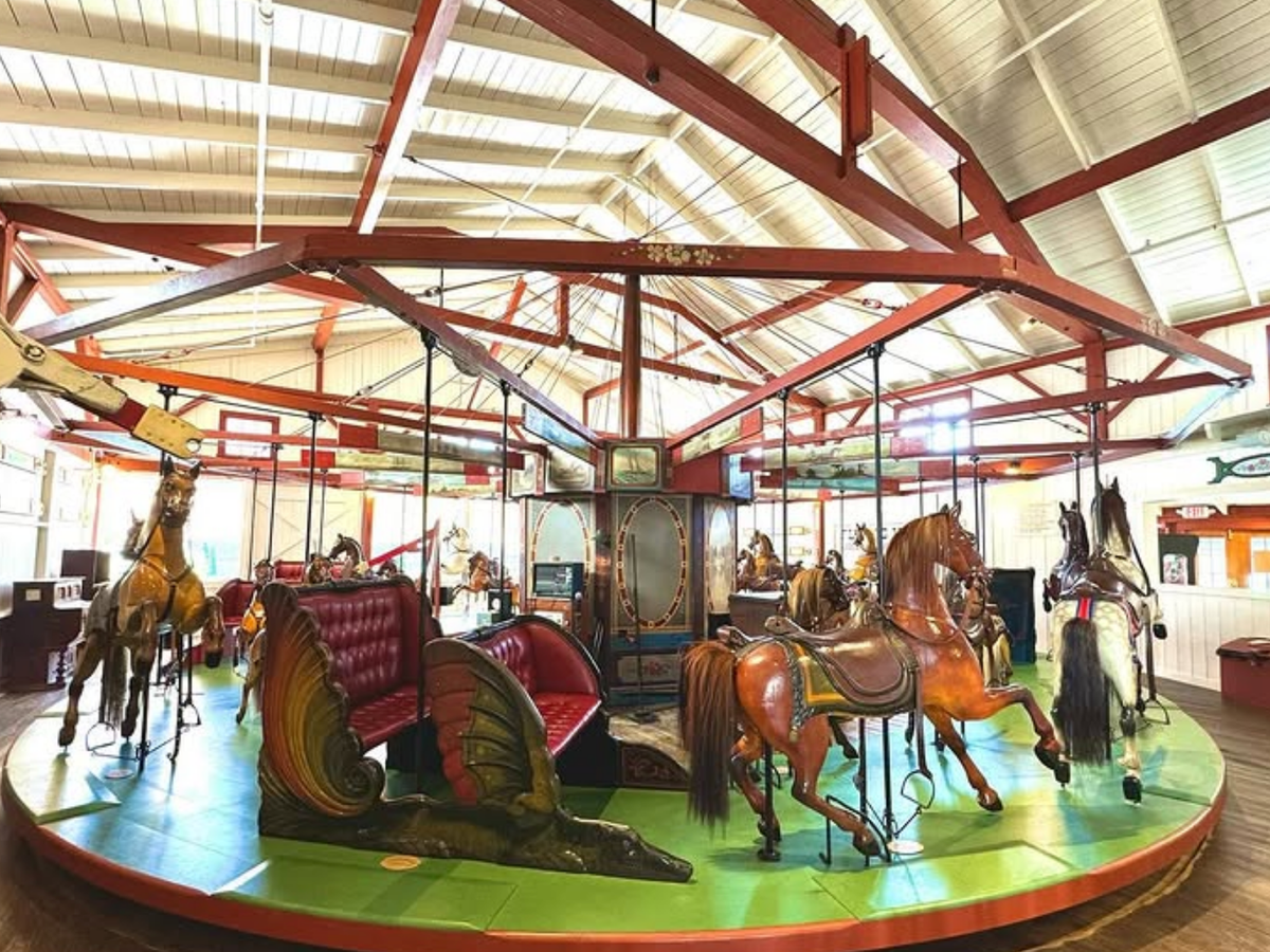 Historic indoor carousel featuring carved wooden horses, colorful benches, and a bright red and white beam ceiling.